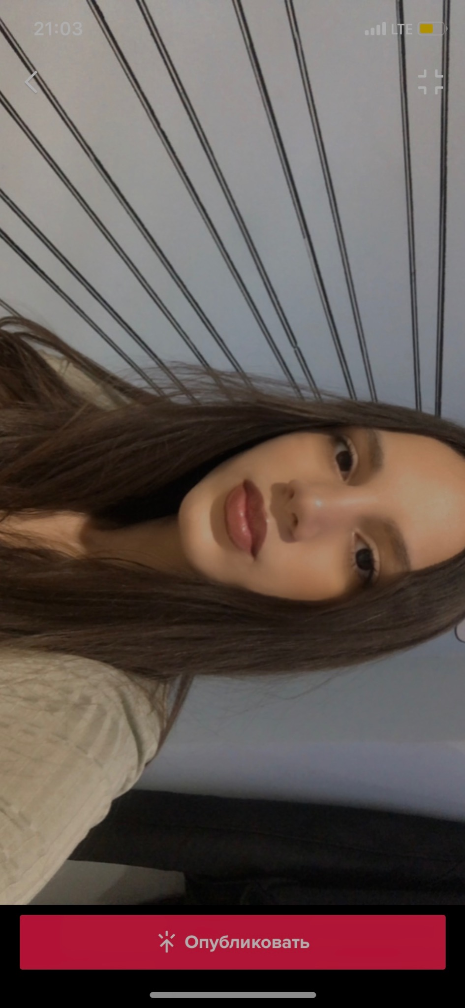 Portret młodej kobiety z brązowymi włosami, skierowanej w stronę obiektywu, z lekkim uśmiechem, na tle ściany z pionowymi, ciemnymi liniami.