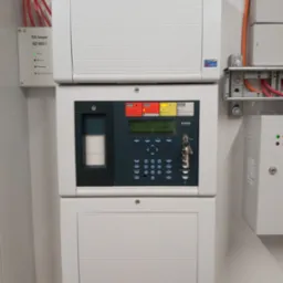 Centrala alarmowa Esser by Honeywell zamontowana na ścianie, z widocznymi kablami i kluczykami w zamku, w tle biała ściana.