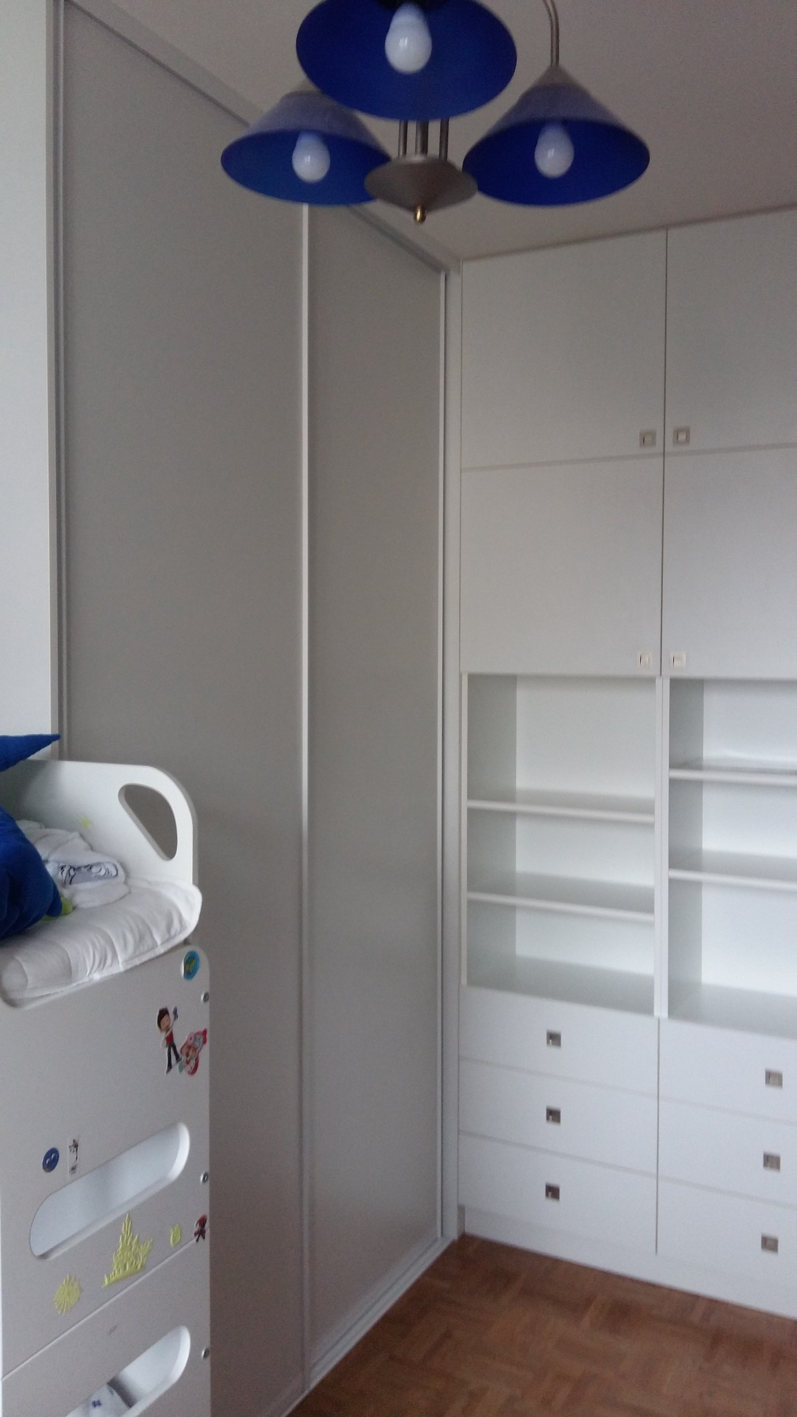 Biała szafa wnękowa z przesuwnymi drzwiami i regałem w pokoju dziecięcym, widoczna część łóżka dziecięcego z naklejkami, oświetlenie sufitowe z trzema niebieskimi kloszami.