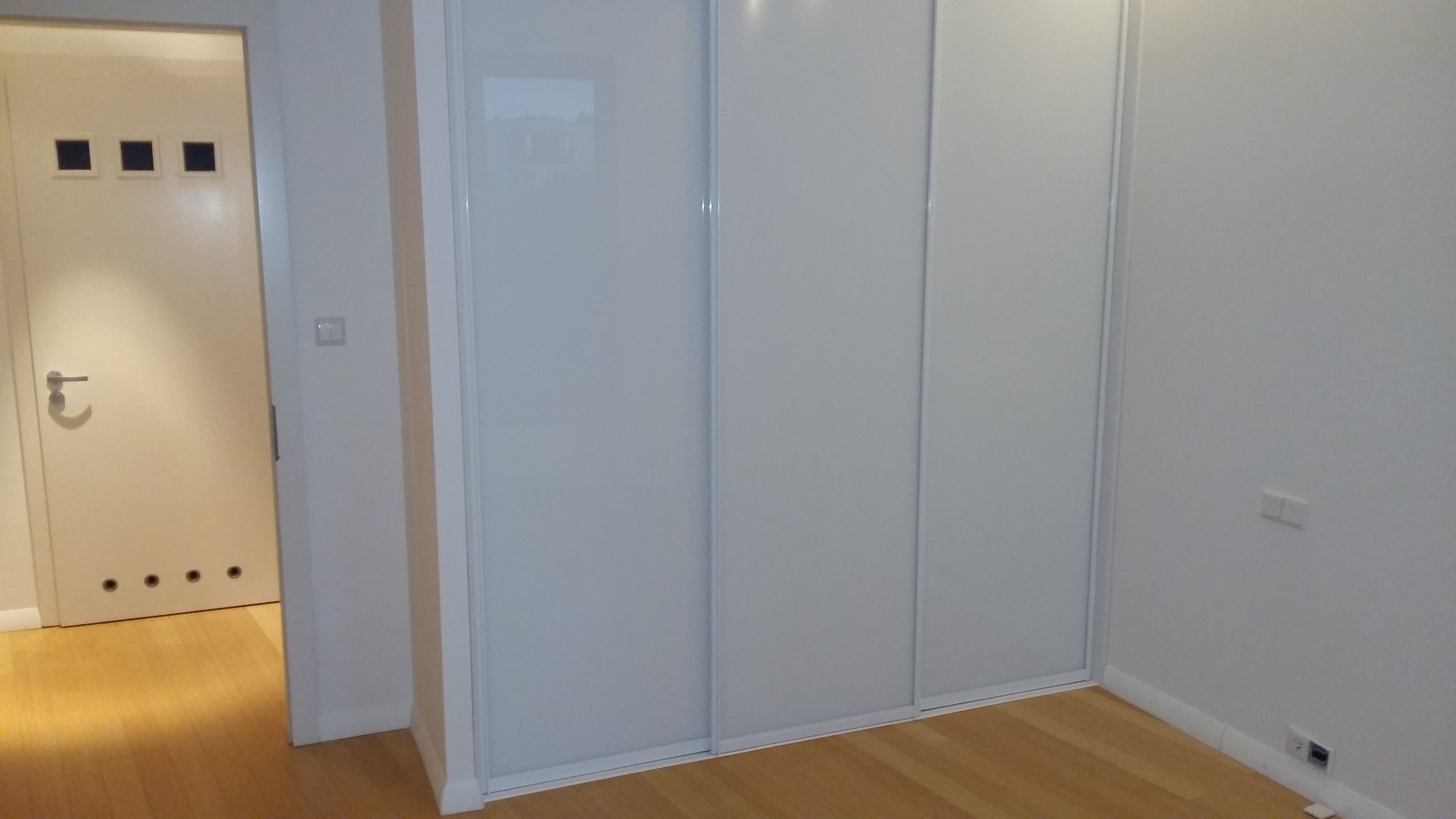 Biała szafa wnękowa z przesuwnymi drzwiami w minimalistycznym wnętrzu, widoczna podłoga z paneli oraz fragment białych drzwi z czarnymi akcentami.