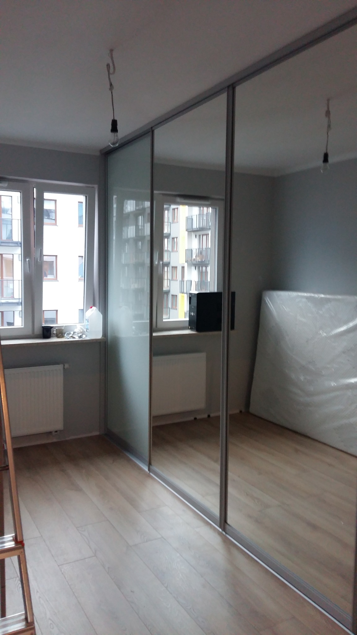 Zamontowana szafa wnękowa z przesuwanymi drzwiami lustrzanymi w pokoju z drewnianą podłogą, widoczne okno z widokiem na budynek mieszkalny, w tle materac owinięty folią.