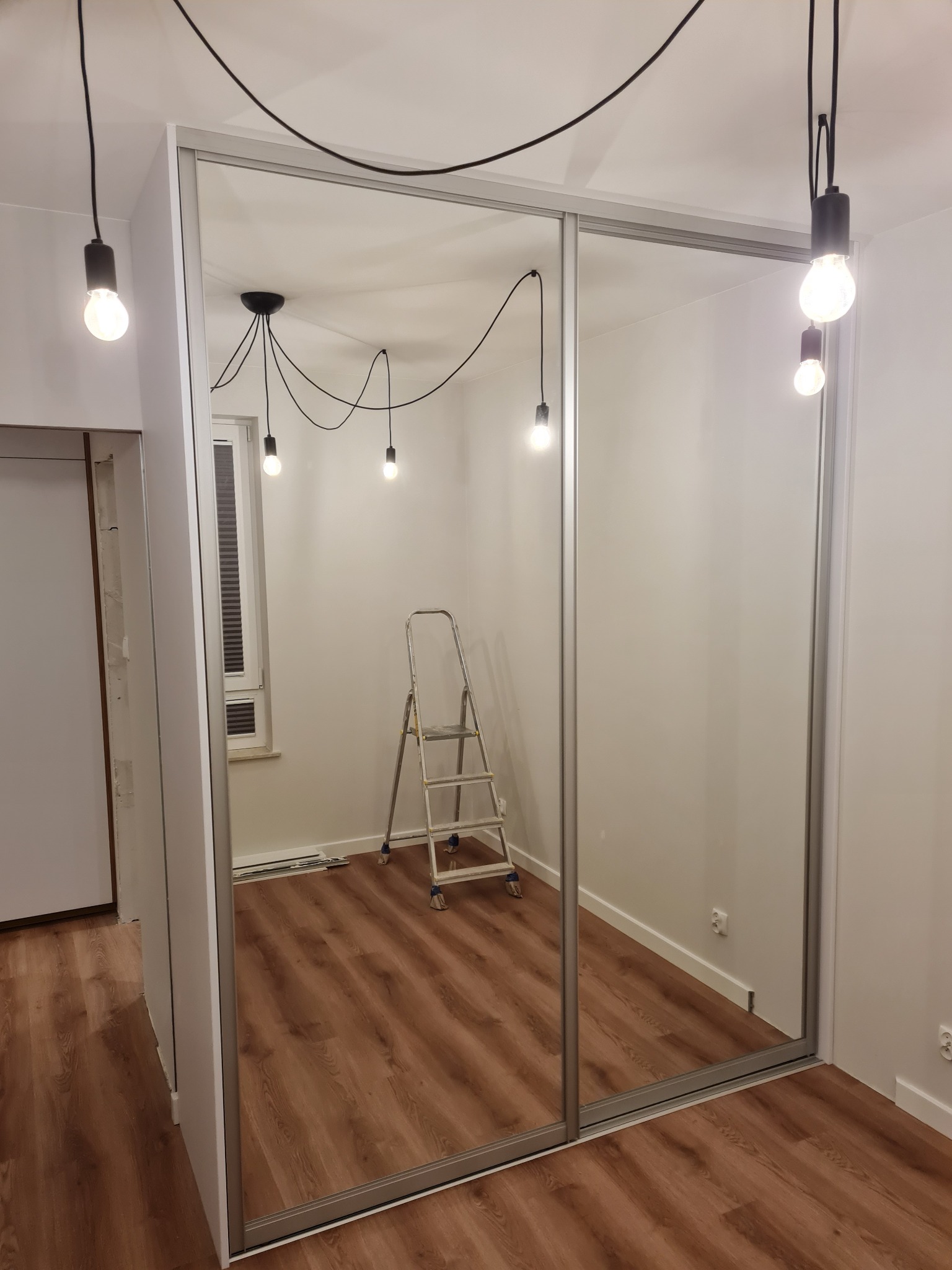 Wnętrze pokoju z zamontowaną szafą wnękową z przesuwnymi, lustrzanymi drzwiami; w tle widoczna drabina malarska i lampy wiszące z żarówkami Edisona.