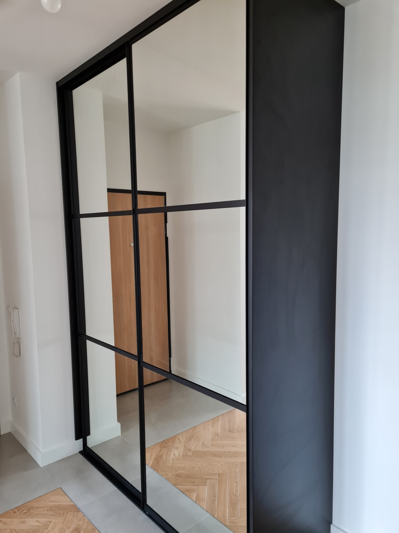 Nowoczesna szafa wnękowa z czarnymi profilami i lustrzanymi drzwiami przesuwnymi, odbijająca fragment korytarza z drewnianą podłogą w jodełkę i białymi ścianami.
