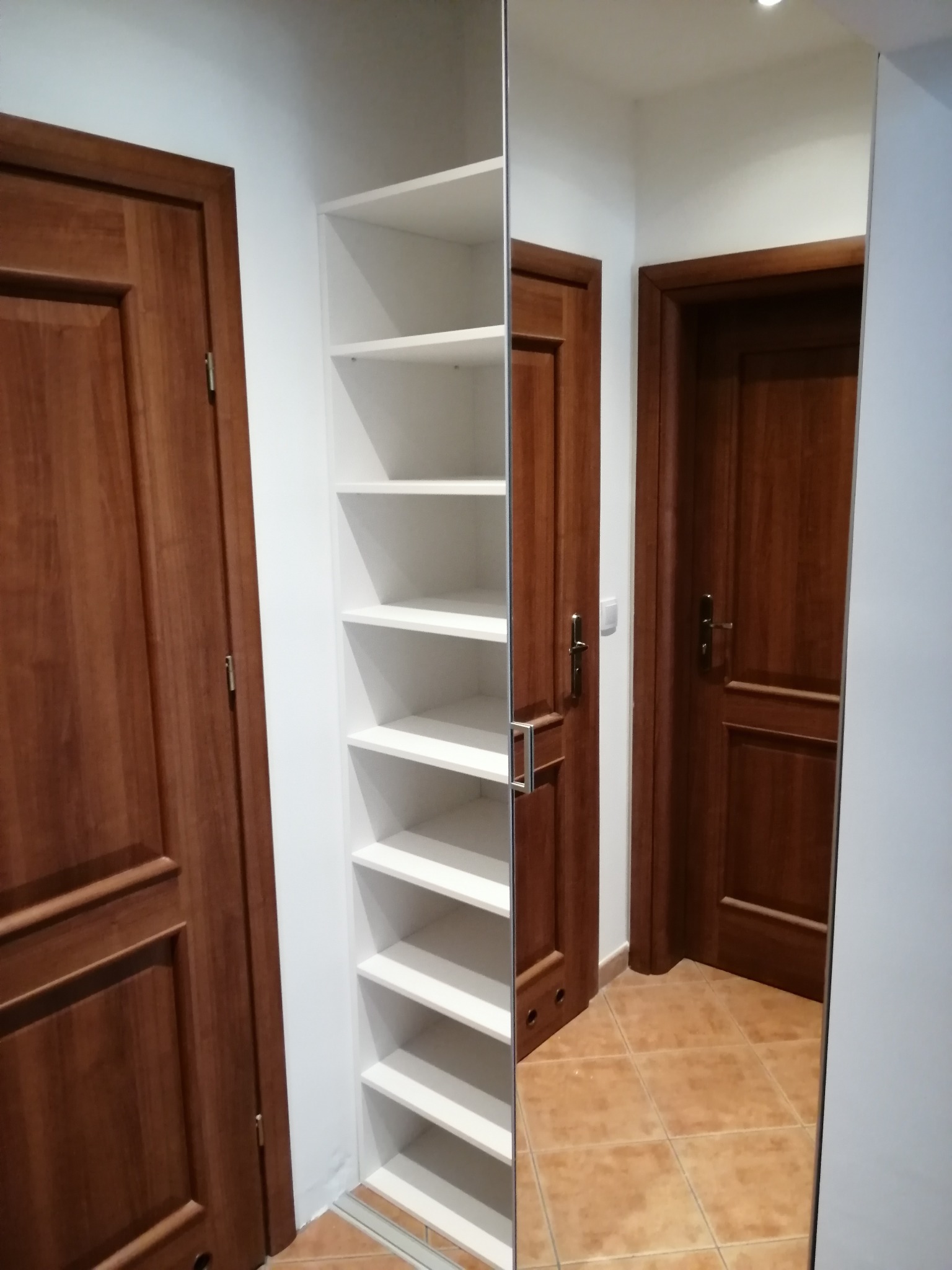 Wąska, biała szafa wnękowa z półkami, umieszczona pomiędzy dwiema drewnianymi drzwiami w przedpokoju z brązową terakotą.