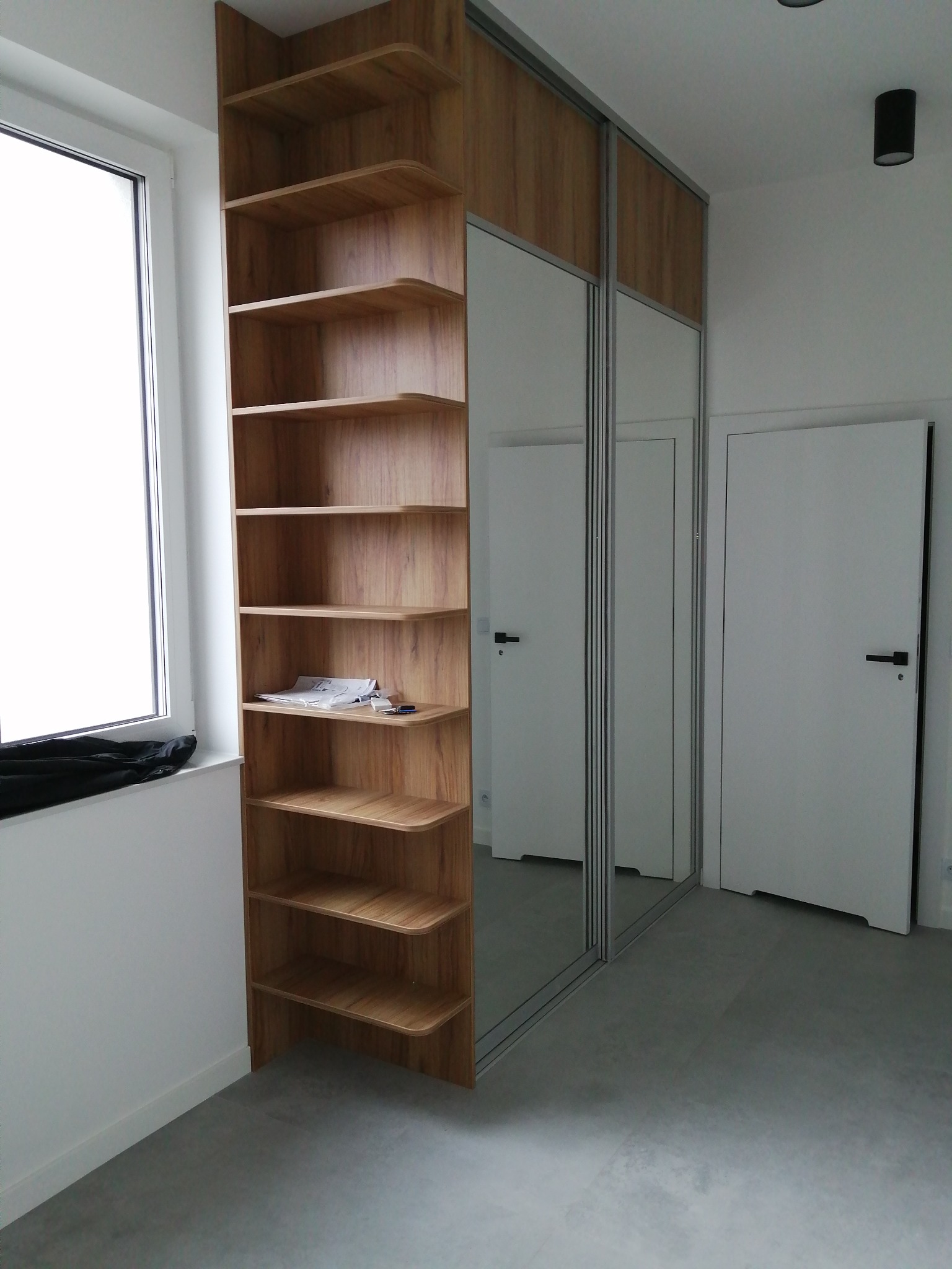 Wnętrze pokoju z szafą wnękową z przesuwnymi drzwiami z lustrami, drewnianym regałem z półkami, białymi drzwiami i szarym betonowym podłożem.