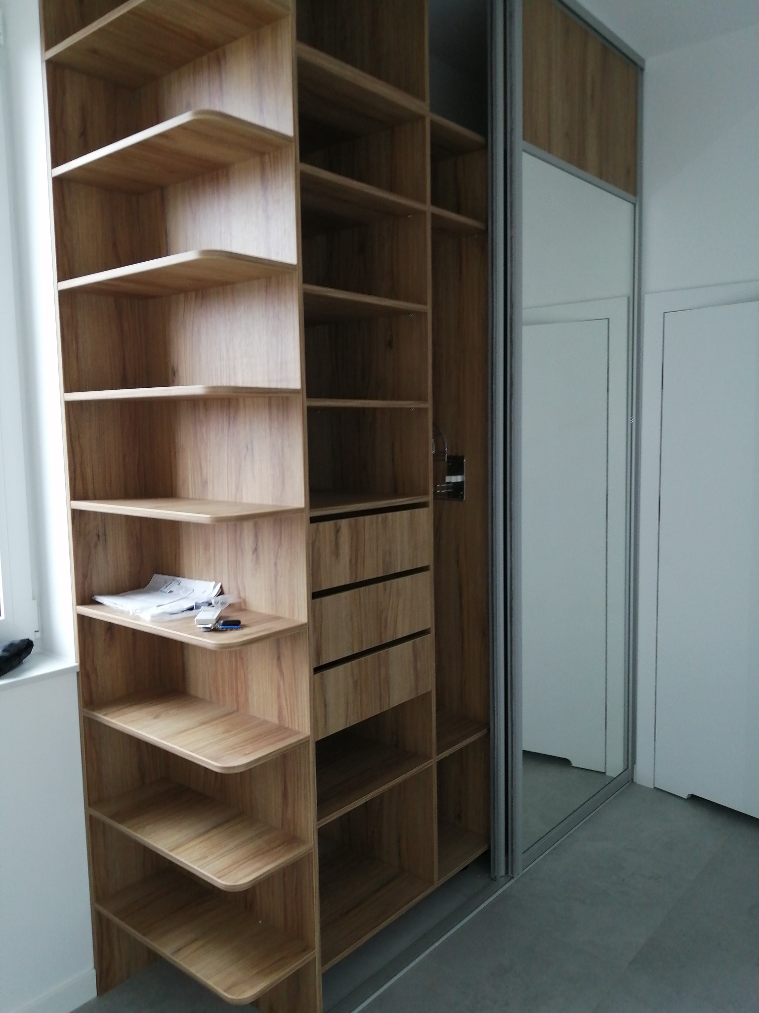 Drewniana szafa wnękowa z przesuwnymi drzwiami lustrzanymi, widoczne półki i szuflady, na jednej z półek leżą dokumenty i klucze.