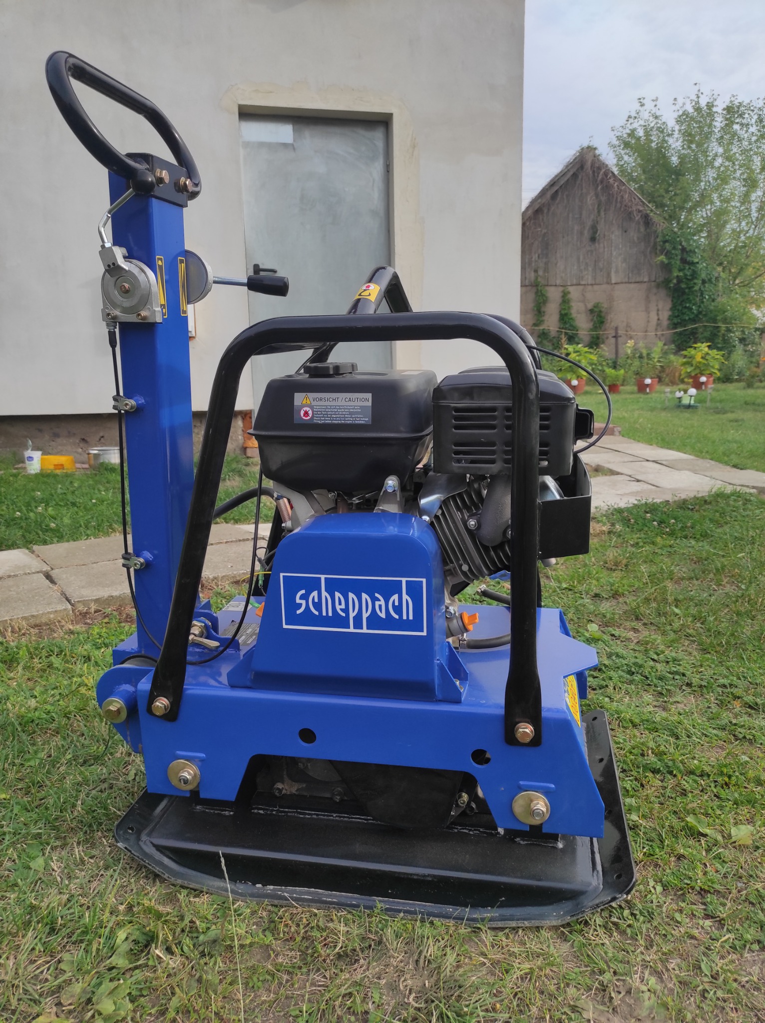 Niebieska zagęszczarka gruntu marki Scheppach, stojąca na trawie przed budynkiem z szarą ścianą i drewnianą stodołą w tle.