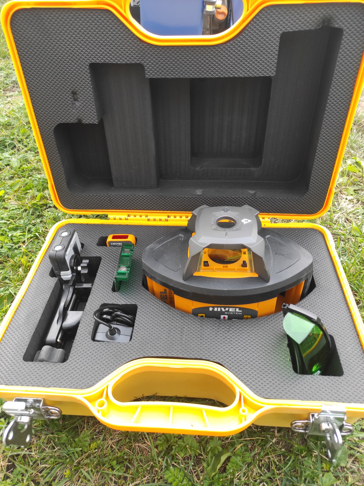 Otwarta żółta walizka transportowa z laserowym niwelatorem obrotowym Nivel, detektorem, łatą i okularami ochronnymi na tle trawy.
