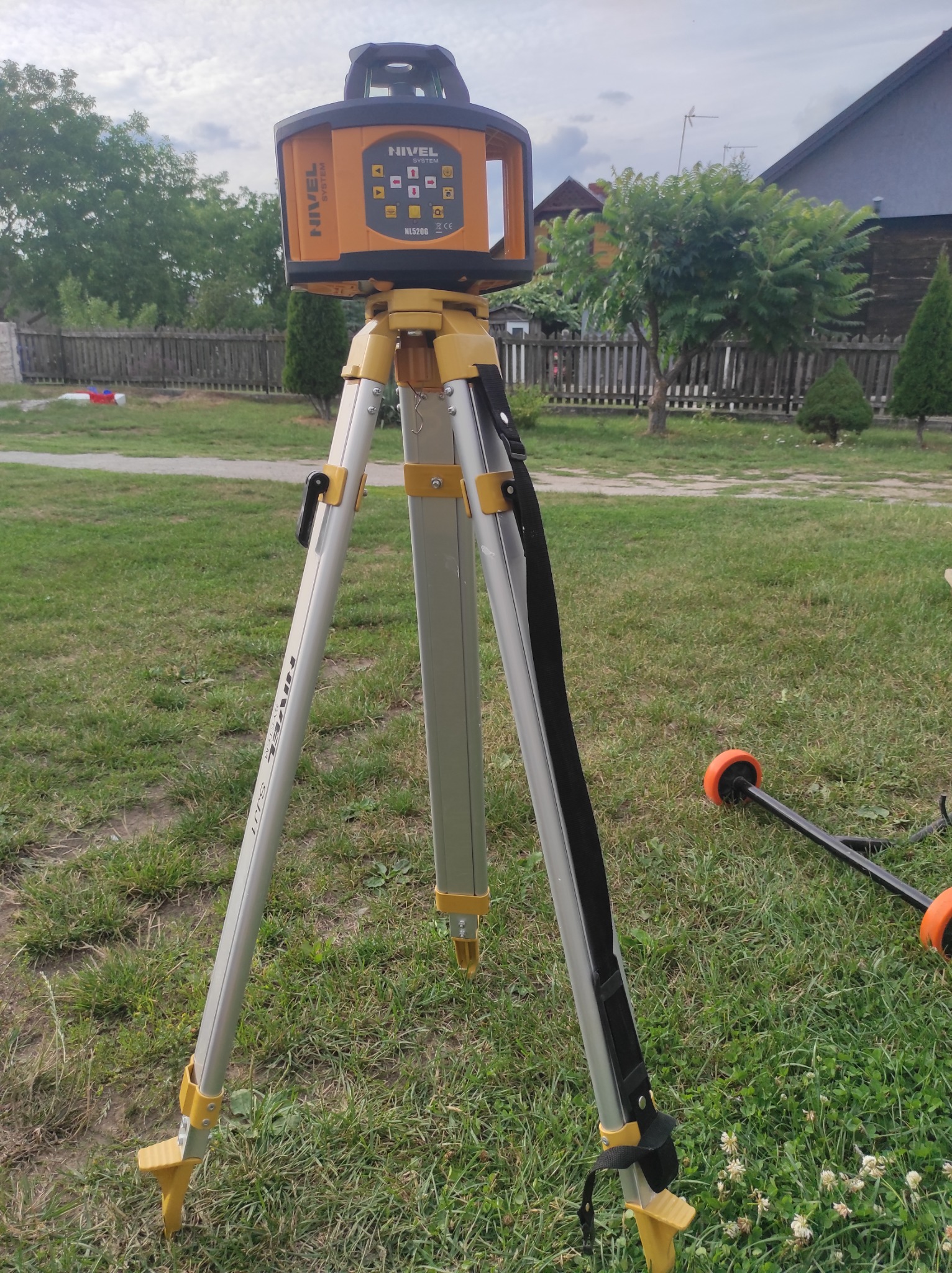 Żółto-pomarańczowy laser krzyżowy Nivel System NL520G na aluminiowym statywie z czarnym paskiem, ustawiony na trawniku przed drewnianym domem i ogrodzeniem.
