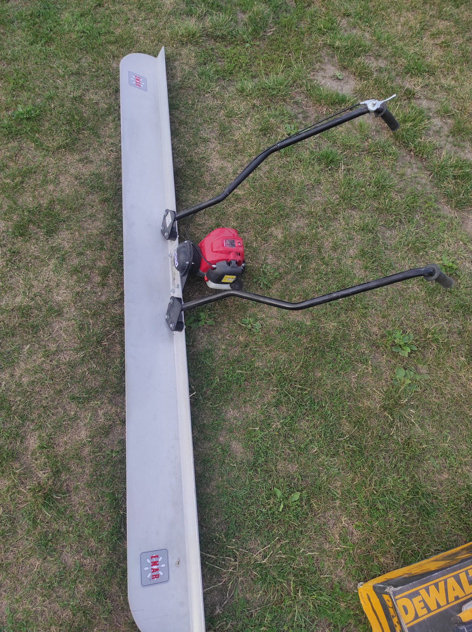 Listwa wibracyjna do betonu z czerwonym silnikiem spalinowym i czarnymi uchwytami, leżąca na trawie obok żółtego pudełka narzędziowego.