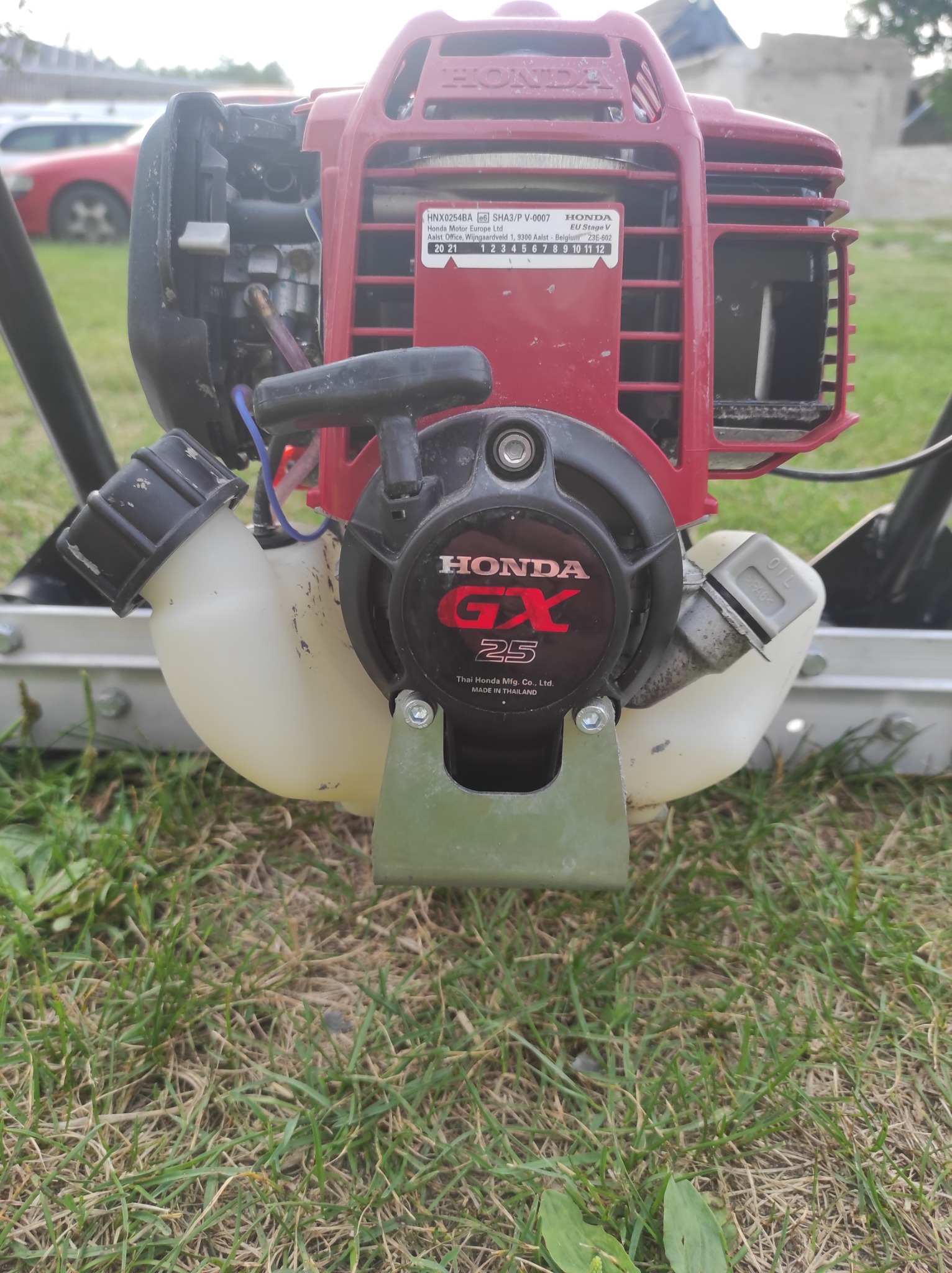 Silnik Honda GX25 zamontowany na wiertnicy glebowej, widok z przodu, na tle trawy i częściowo widocznego samochodu.