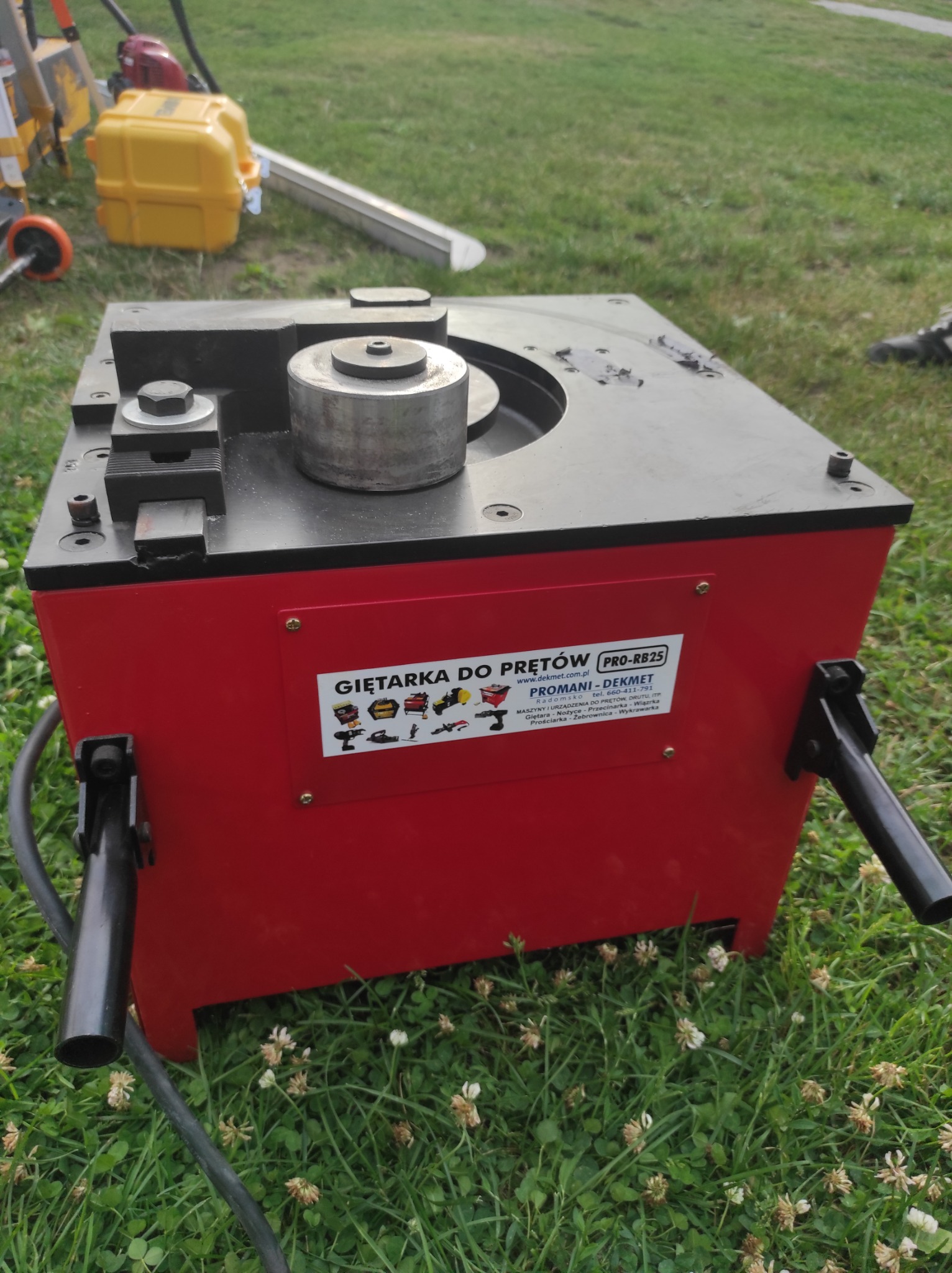 Czerwona giętarka do prętów PRO-RB25 marki Promani-Dekmet stoi na trawie, widoczne elementy robocze i naklejka z danymi producenta.