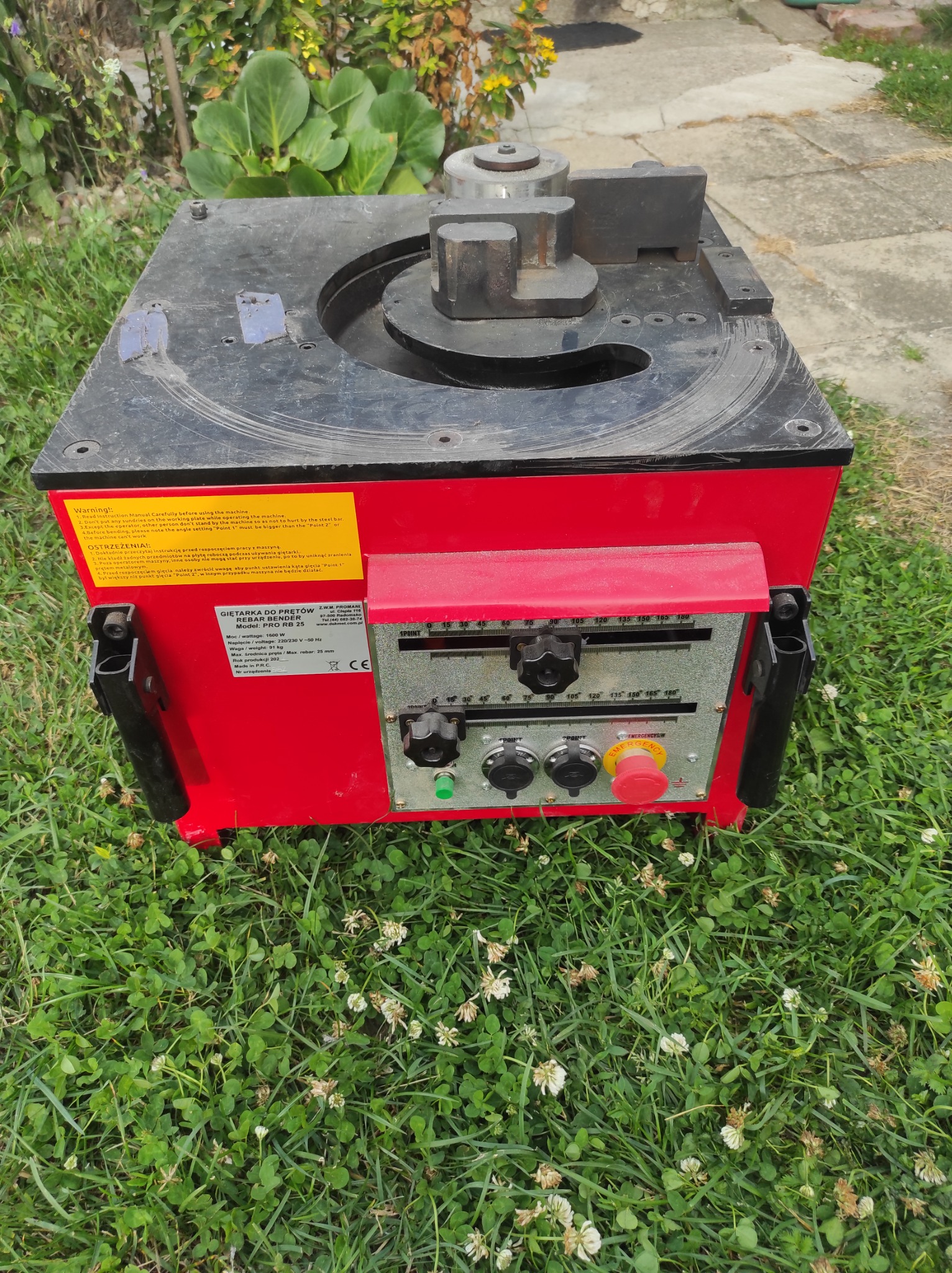 Giętarka do prętów PRO RB 25 na trawie, z widocznymi elementami sterującymi i ostrzeżeniami na czerwonej obudowie.