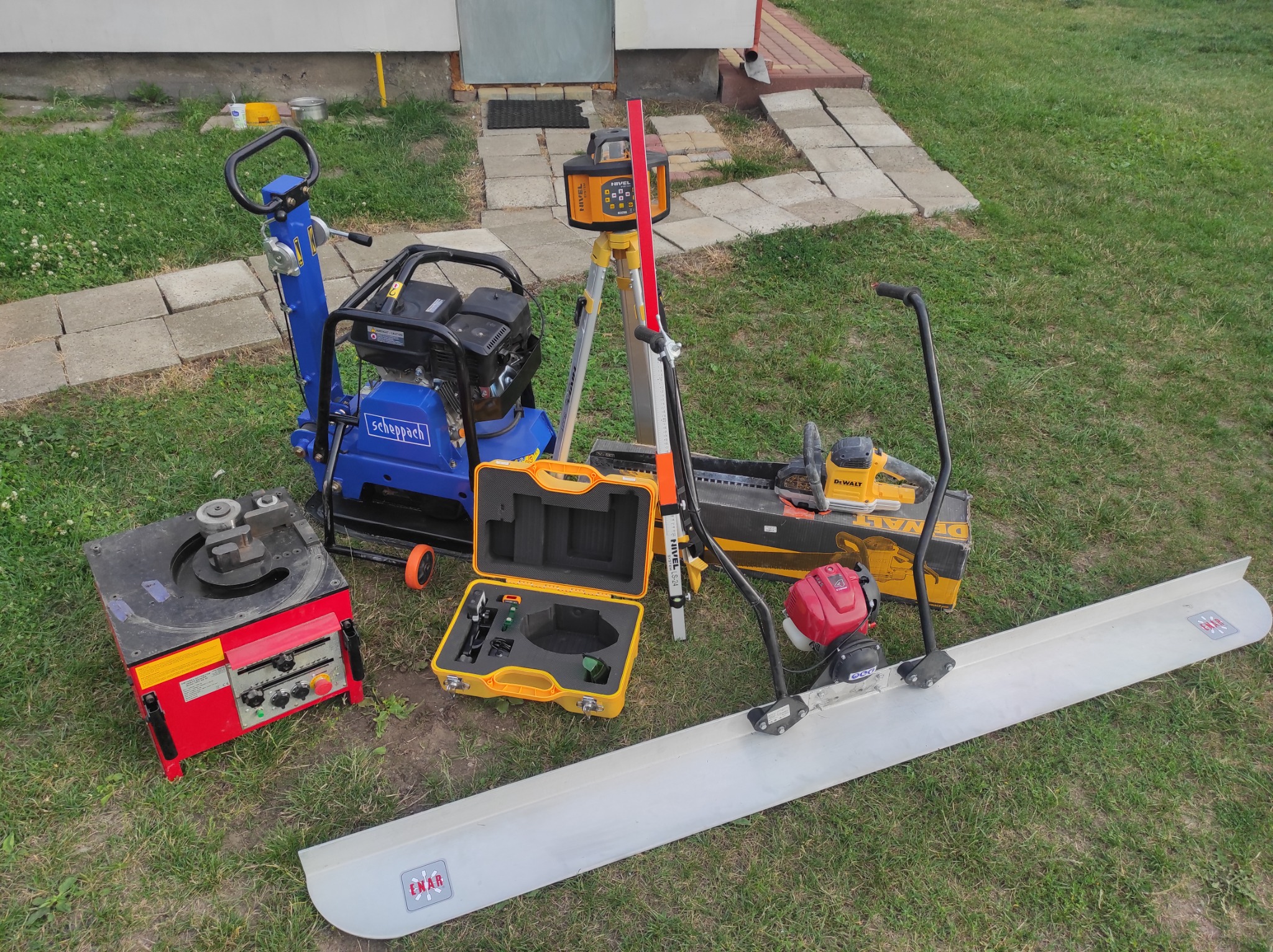 Zestaw sprzętu budowlanego na trawie: zagęszczarka gruntu Scheppach, giętarka do prętów, niwelator laserowy Nivel System na statywie, listwa wibracyjna Enar i szlifierka do betonu DeWalt w kartonie.