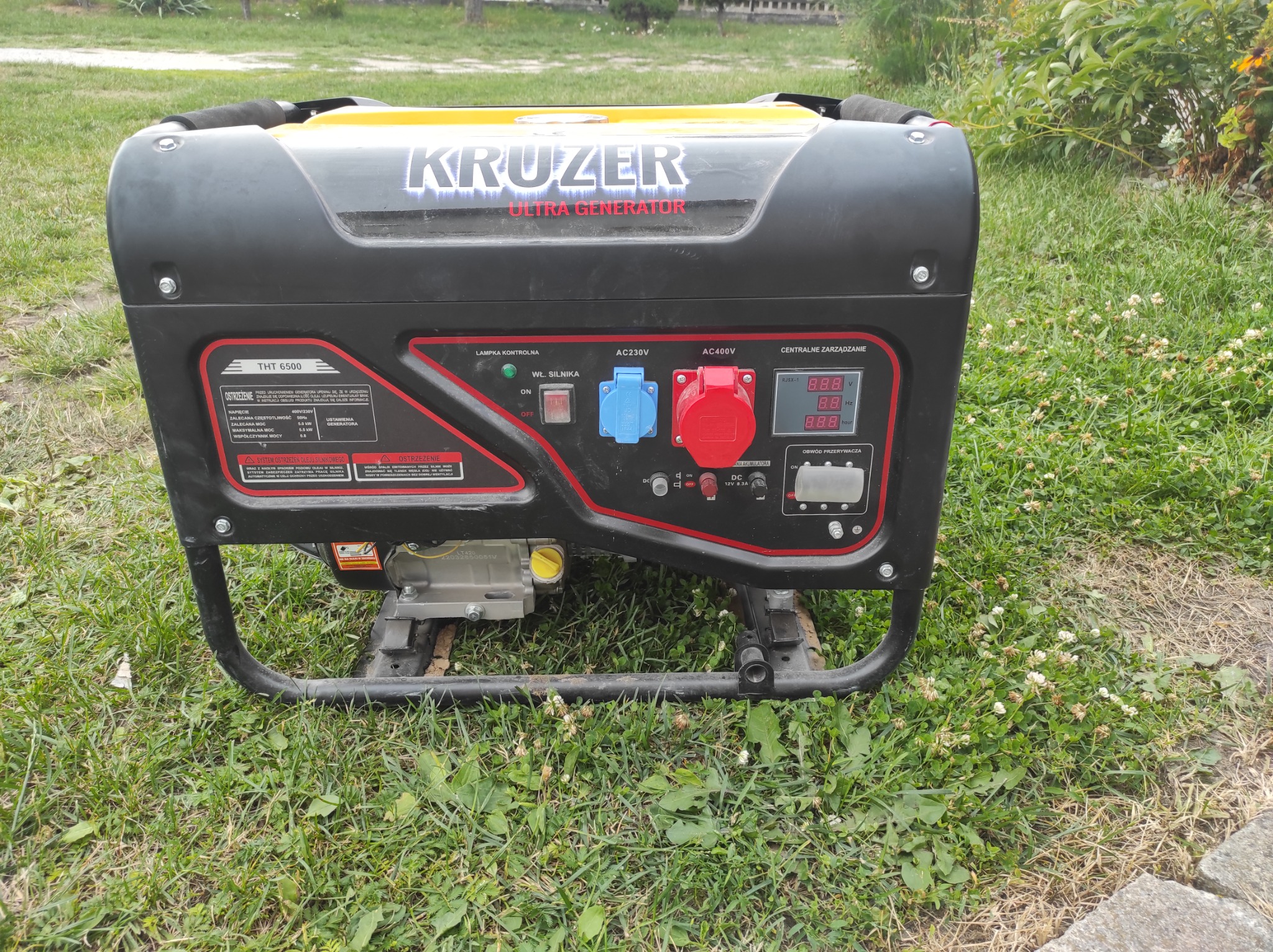 Agregat prądotwórczy Kruzer Ultra Generator THT 6500 na trawie, widoczne gniazda AC 230V i 400V, panel sterowania z wyświetlaczami oraz ostrzeżenia dotyczące oleju silnikowego i obsługi.