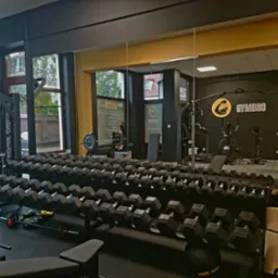 Wnętrze siłowni z rzędem hantli, maszynami do ćwiczeń i lustrem odzwierciedlającym logo 'GYMBRO' na ścianie.