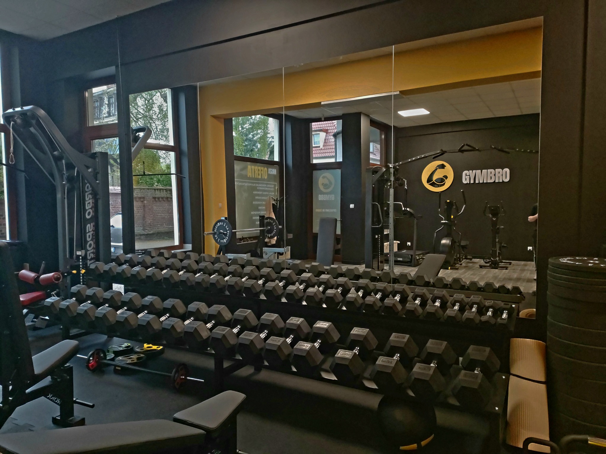 Wnętrze siłowni z rzędem hantli, maszynami do ćwiczeń i lustrem odzwierciedlającym logo 'GYMBRO' na ścianie.