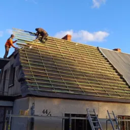 Ekipa dekarzy montuje drewnianą konstrukcję dachu na dwuspadowym budynku z częściowo wymienionym pokryciem z blachy falistej, widoczne okna dachowe i antena telewizyjna.