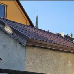 Brązowe blaszane pokrycie dachu na starym domu z widoczną wieżą kościoła w tle.