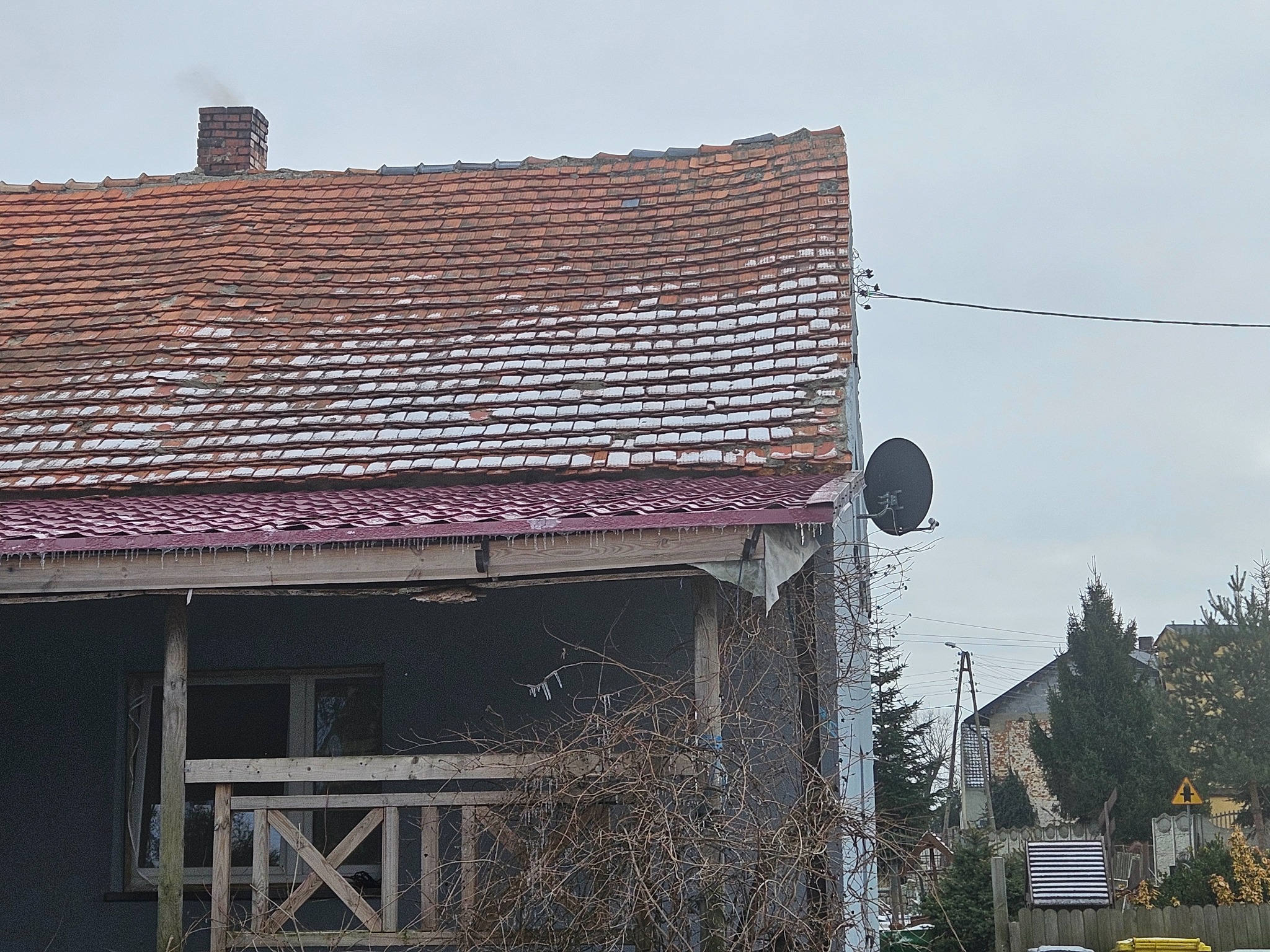 Dom z widocznym, częściowo uszkodzonym dachem z czerwonej dachówki, pokrytym miejscami śniegiem, z kominem, anteną satelitarną i soplami lodu zwisającymi z okapu