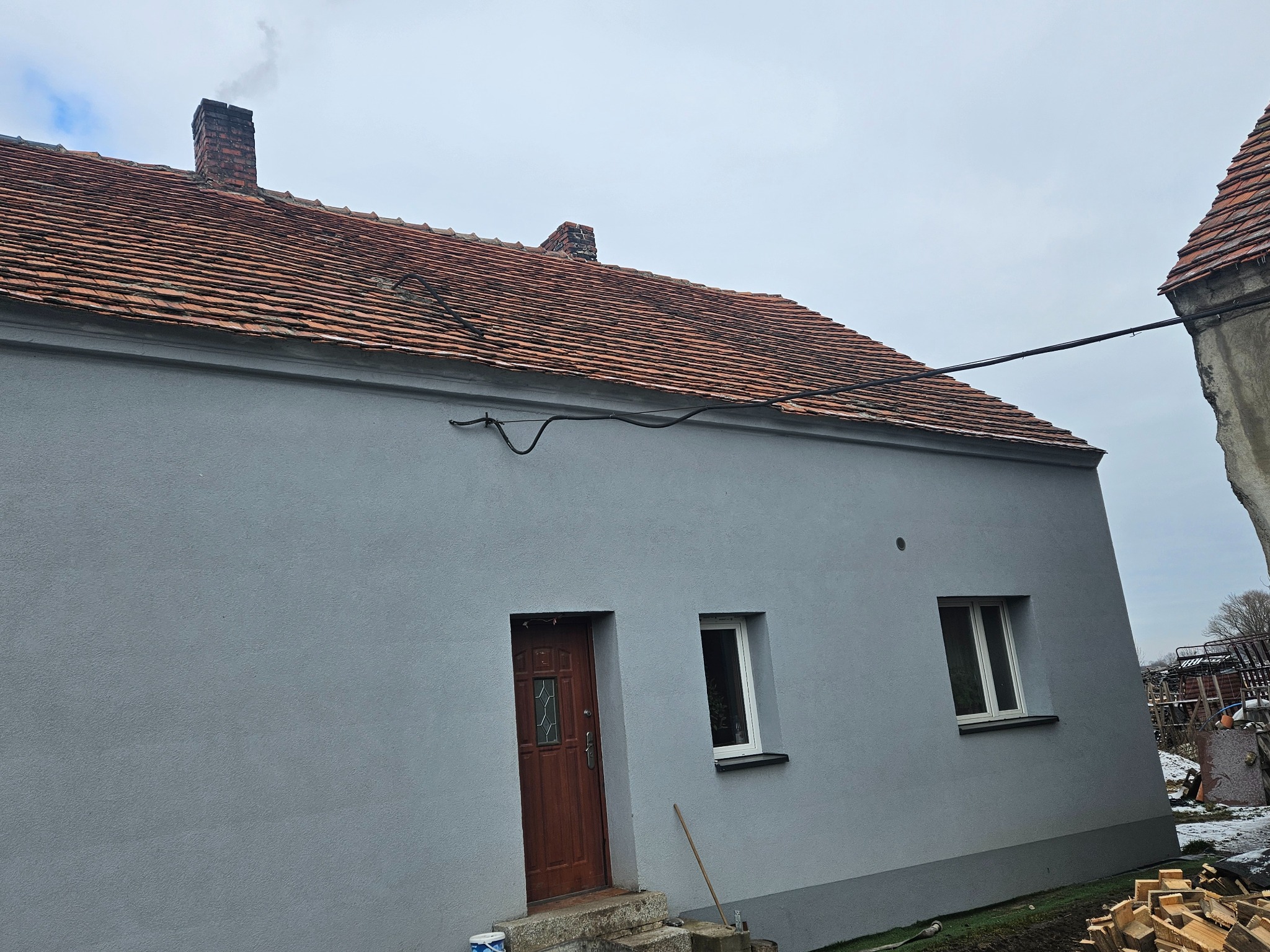 Dom jednorodzinny z nową, czerwoną dachówką ceramiczną, widoczny komin z cegły i świeżo otynkowana elewacja w odcieniach szarości. Na pierwszym planie stos drewna i fragment sąsiedniego budynku.
