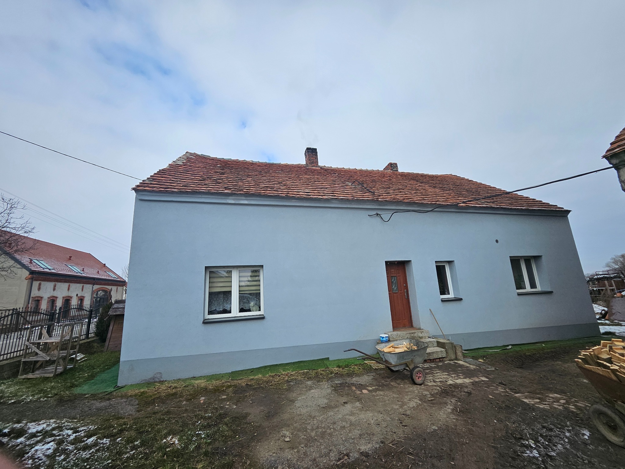 Dom jednorodzinny w Lubinie z ceramiczną dachówką, przygotowany do prac remontowych, wózki z drewnem i materiałami budowlanymi przed wejściem.