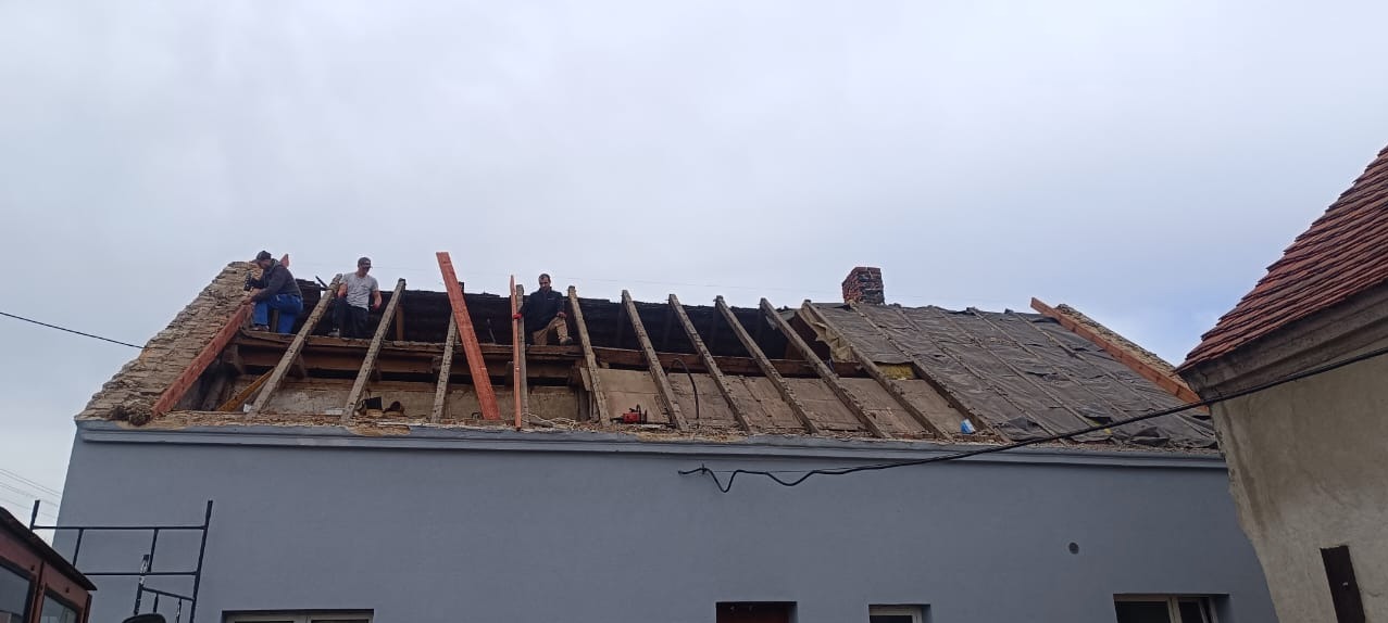 Ekipa dekarzy pracuje na częściowo zdemontowanym dachu starego domu; widoczne krokwie, fragment starego pokrycia, komin i budynek sąsiada z czerwoną dachówką.