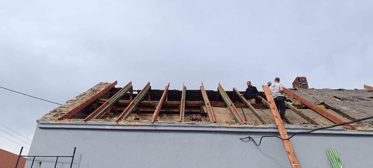 Demontaż starego dachu z widocznymi elementami konstrukcyjnymi, pracownicy na dachu, częściowo zdjęte pokrycie, komin w tle.