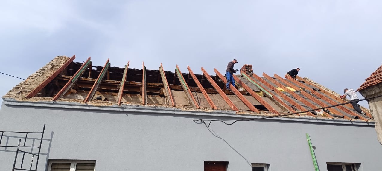 Ekipa dekarzy demontuje starą konstrukcję dachu z ceglanego domu, widoczne krokwie i elementy starego poszycia, jeden z pracowników stoi na dachu obok komina, drugi pracuje w oddali.