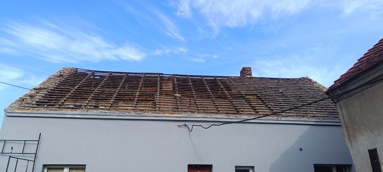 Uszkodzony dach z widoczną konstrukcją drewnianą i fragmentami starej dachówki, z kominem w tle, na tle błękitnego nieba z chmurami.