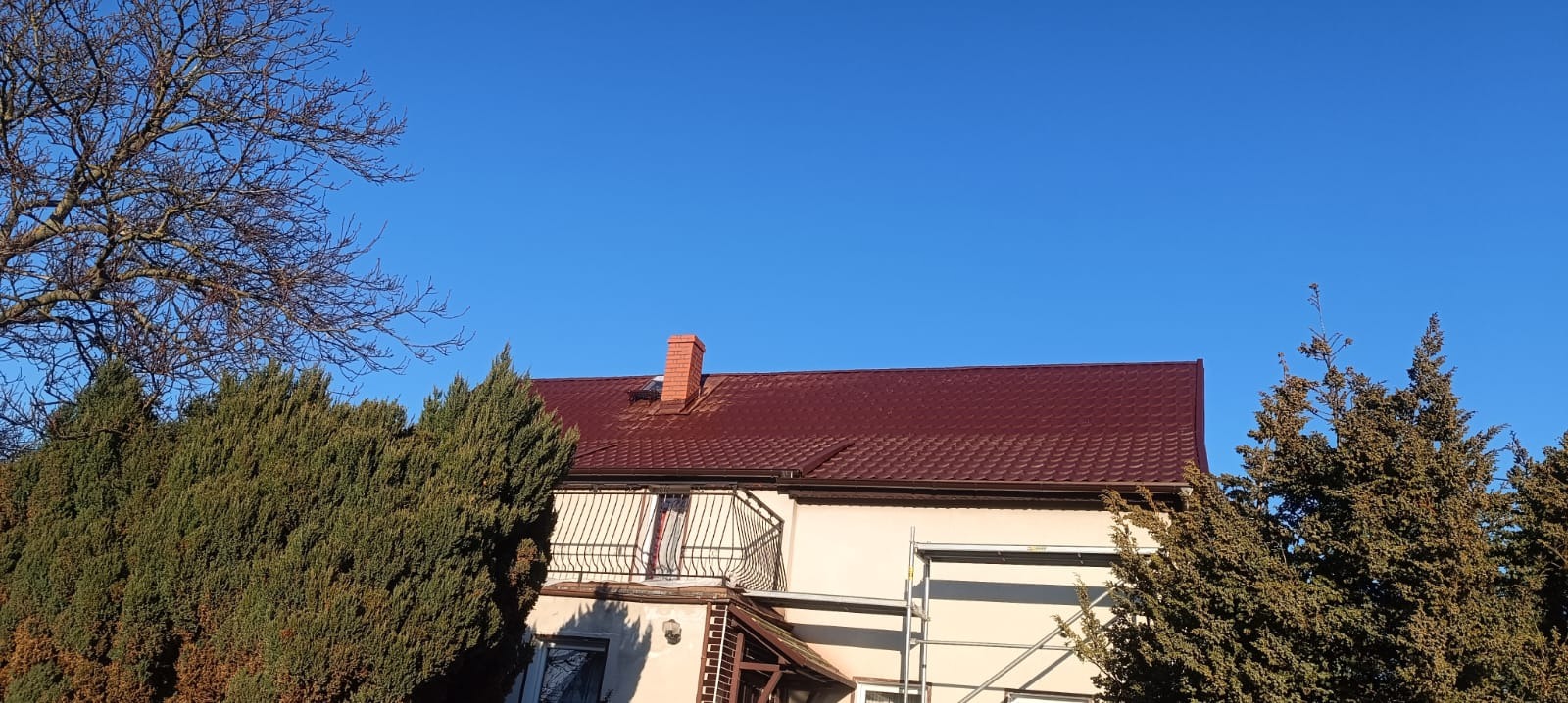 Dom jednorodzinny z nowym, bordowym dachem z blachodachówki trapezowej, widoczne rusztowanie przy ścianie bocznej i ceglany komin