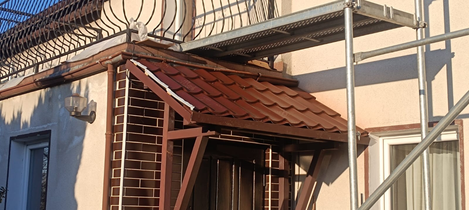 Zadaszenie nad wejściem do budynku z brązową dachówką, widoczne rusztowanie i balkon z metalową balustradą.