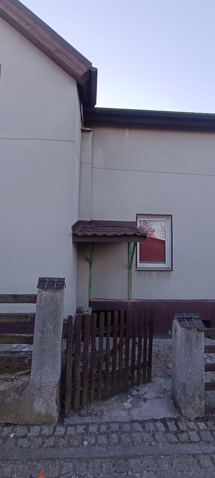 Fragment elewacji budynku z brązowym dachem, drewnianą podbitką i małym zadaszeniem nad wejściem wspartym na zielonych słupkach. Widoczny fragment drewnianego płotu i betonowe słupki.