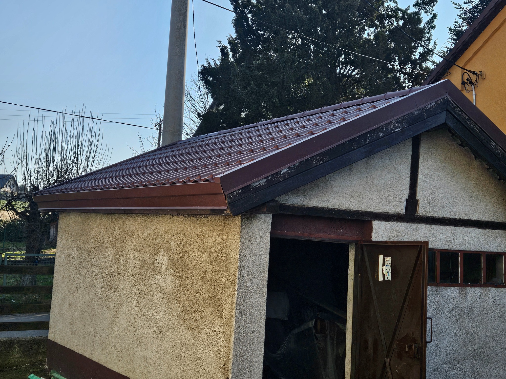 Brązowy dach z blachy trapezowej na małym budynku gospodarczym z otwartymi drzwiami, widoczny słup energetyczny i drzewa w tle.