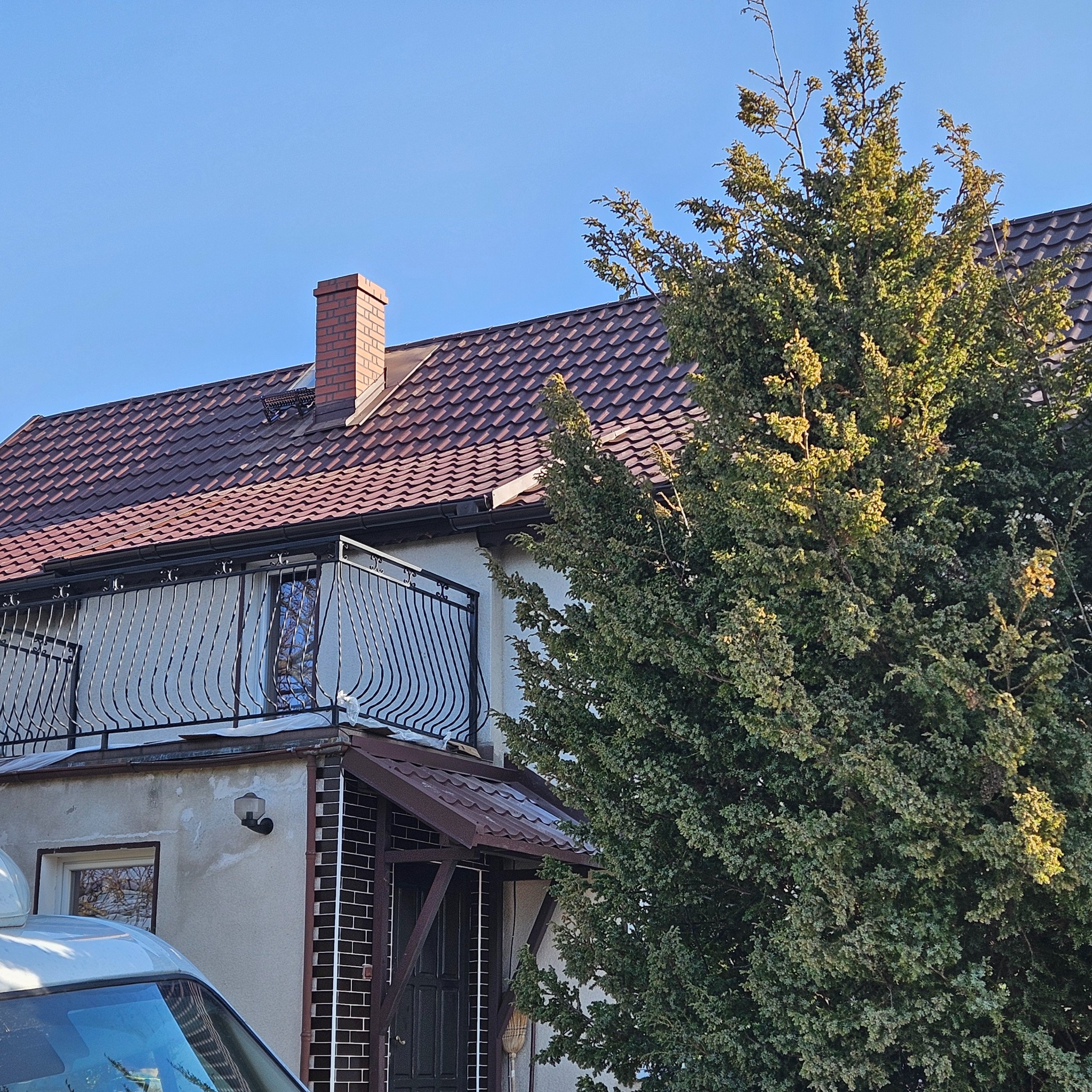 Dom jednorodzinny z nową, brązową dachówką ceramiczną i ceglanym kominem, widoczne także balustrady balkonu i duży iglak.