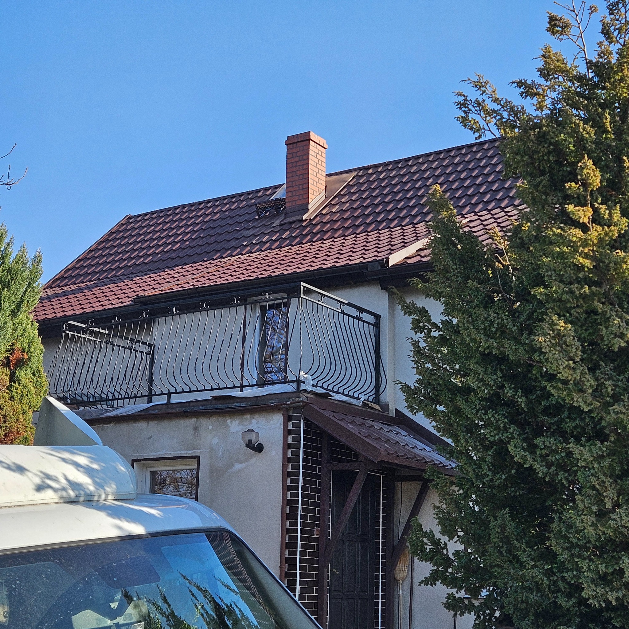 Dom z nowym brązowym dachem z dachówki ceramicznej, ceglanym kominem i balkonem z ozdobną, czarną balustradą, częściowo zasłonięty przez drzewa i fragment samochodu dostawczego.
