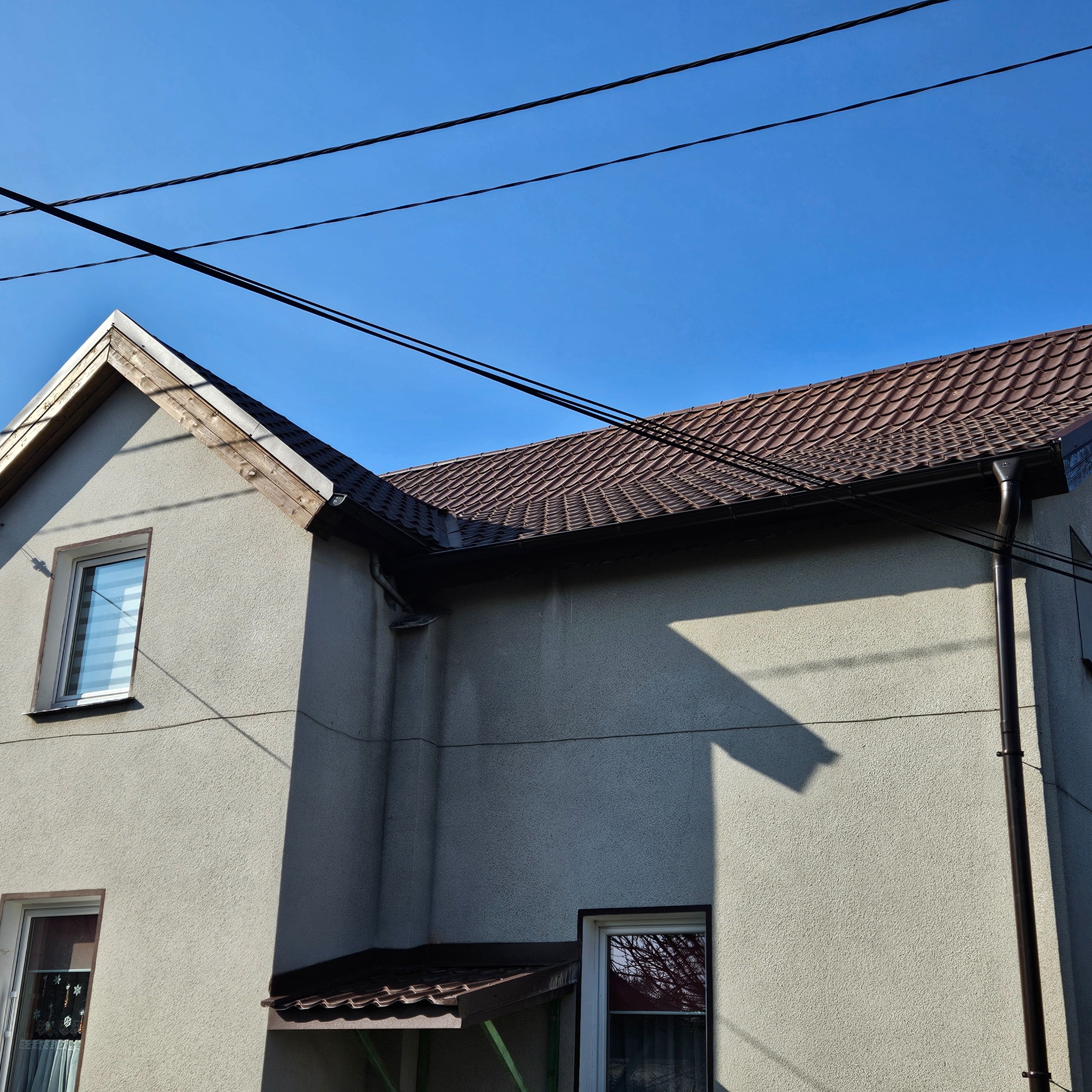 Dom z nowym, brązowym dachem z dachówki ceramicznej, rynny w kolorze dachu, widoczne okna i elewacja w jasnym kolorze, na tle błękitnego nieba i przewodów elektrycznych.