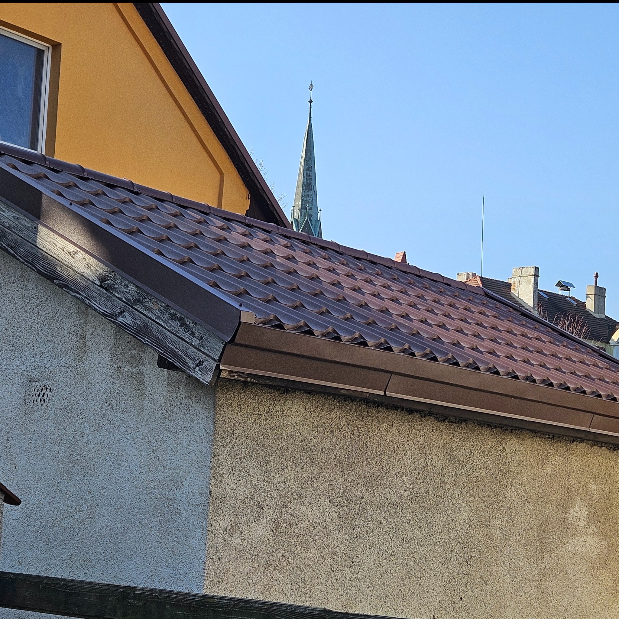 Brązowe blaszane pokrycie dachu na starym domu z widoczną wieżą kościoła w tle.