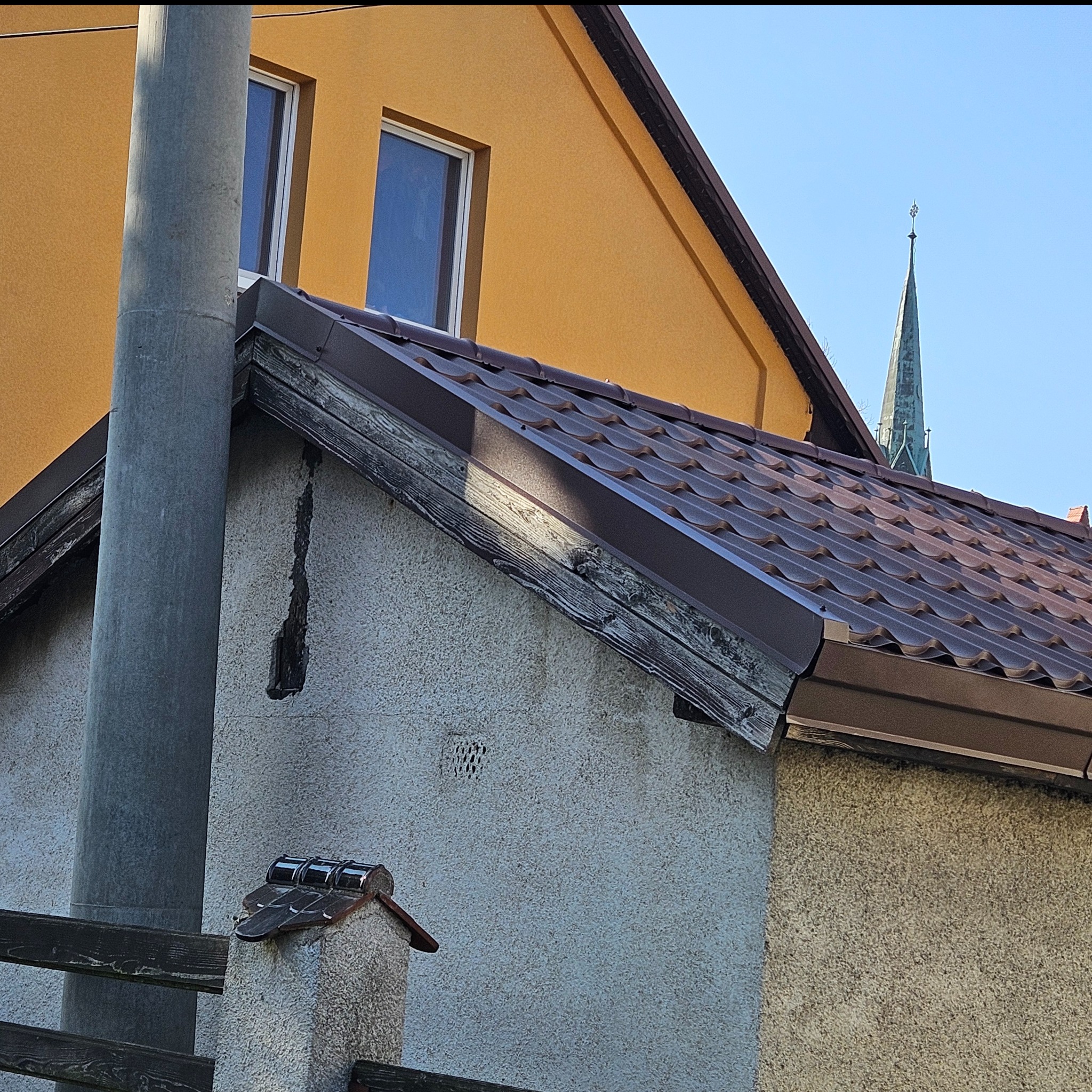 Brązowa blachodachówka na szczycie budynku z widoczną starą konstrukcją drewnianą, pęknięciem w ścianie, słupem elektrycznym i iglicą kościoła w tle.