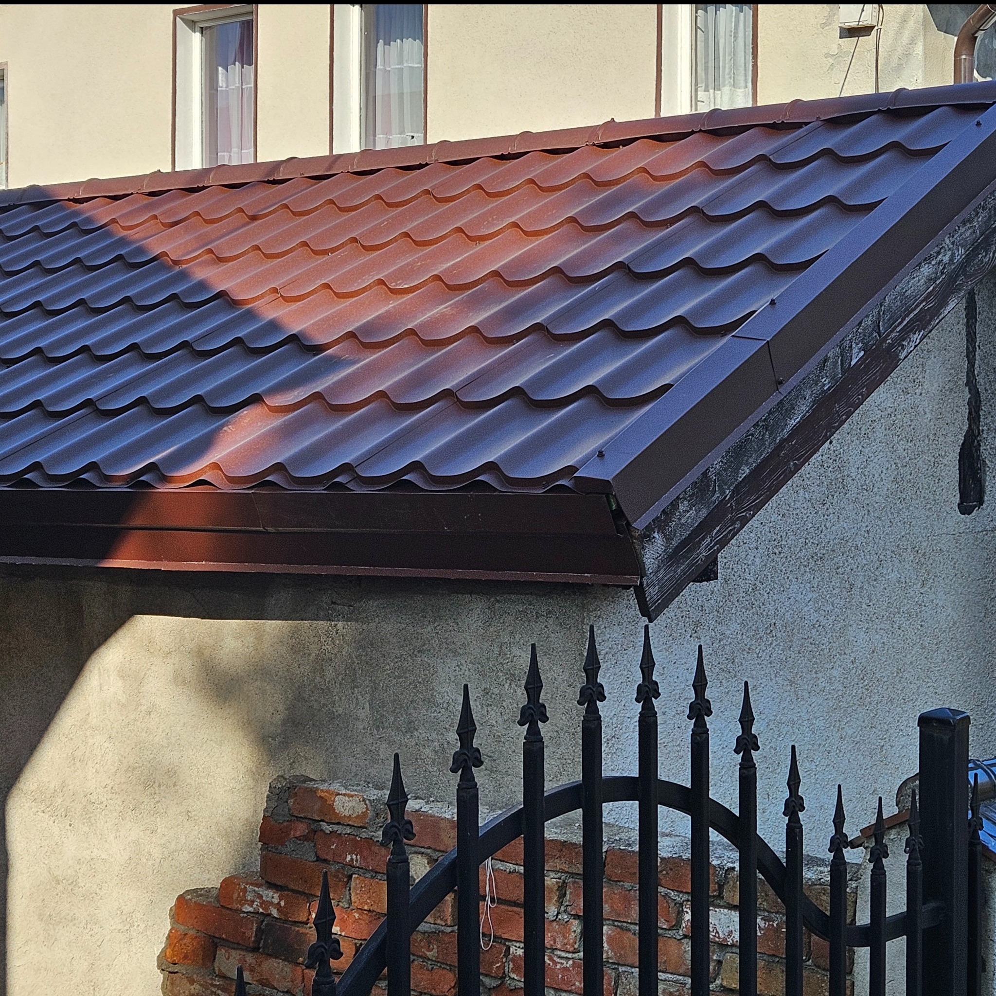 Metalowa dachówka w kolorze ciemnej czekolady na domu z oknami, widziana zza czarnego, kutego ogrodzenia.