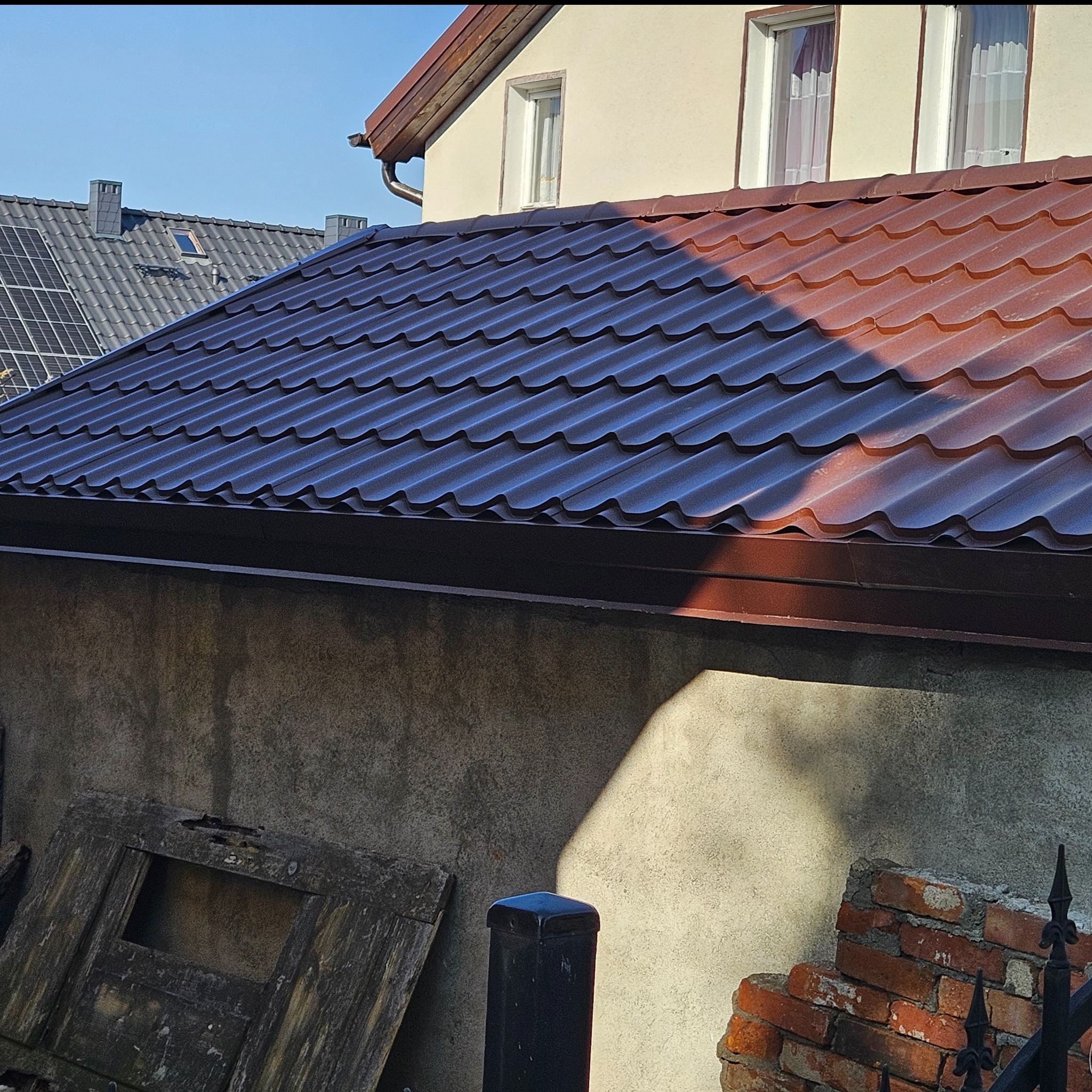 Nowe, brązowe pokrycie dachowe z blachy trapezowej, widoczne z dołu, z fragmentem elewacji budynku i elementami starego muru w tle.