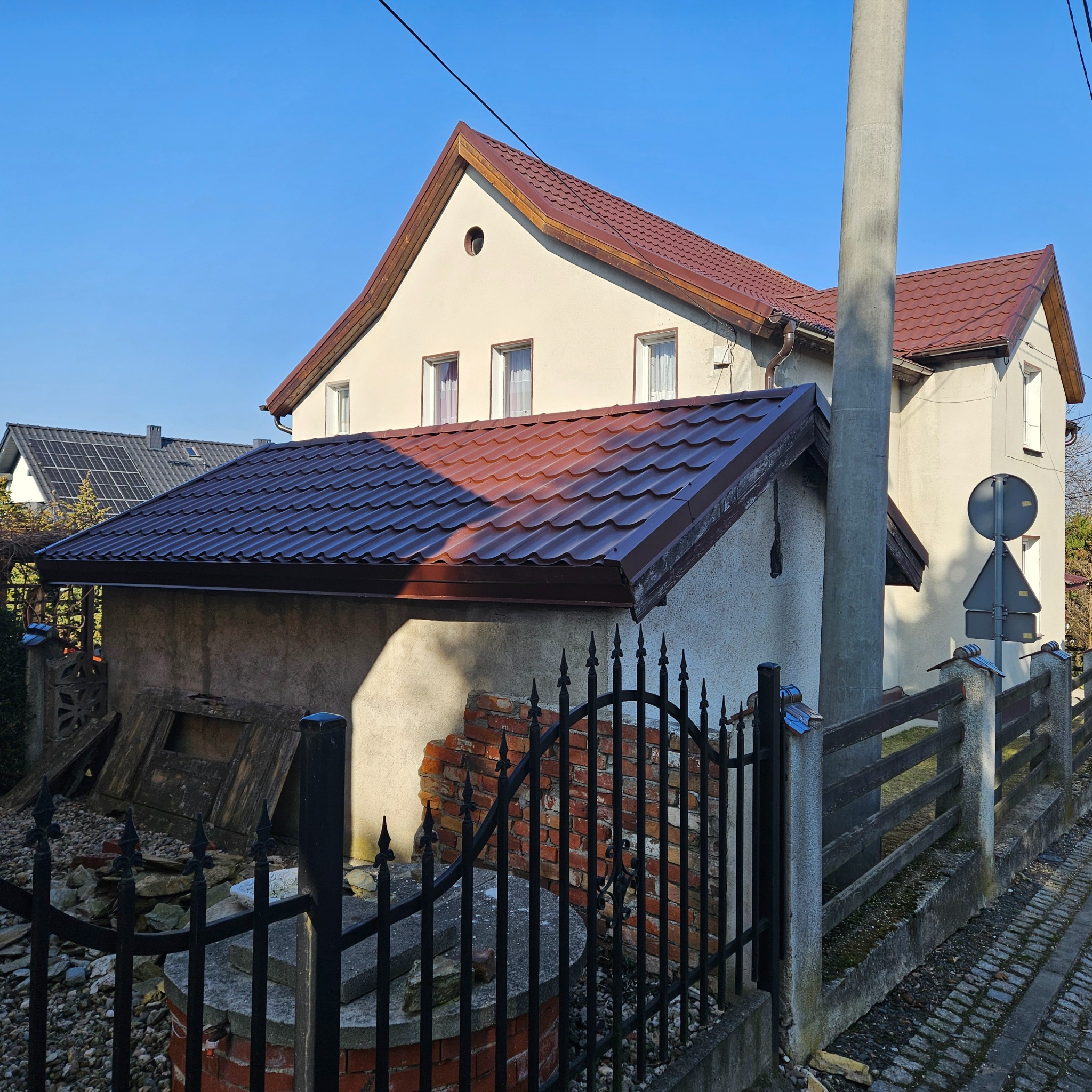Dom jednorodzinny z nowym, brązowym dachem z blachodachówki, widoczny z perspektywy ulicy, z fragmentem zniszczonego muru i czarnym, kutym ogrodzeniem na pierwszym planie.
