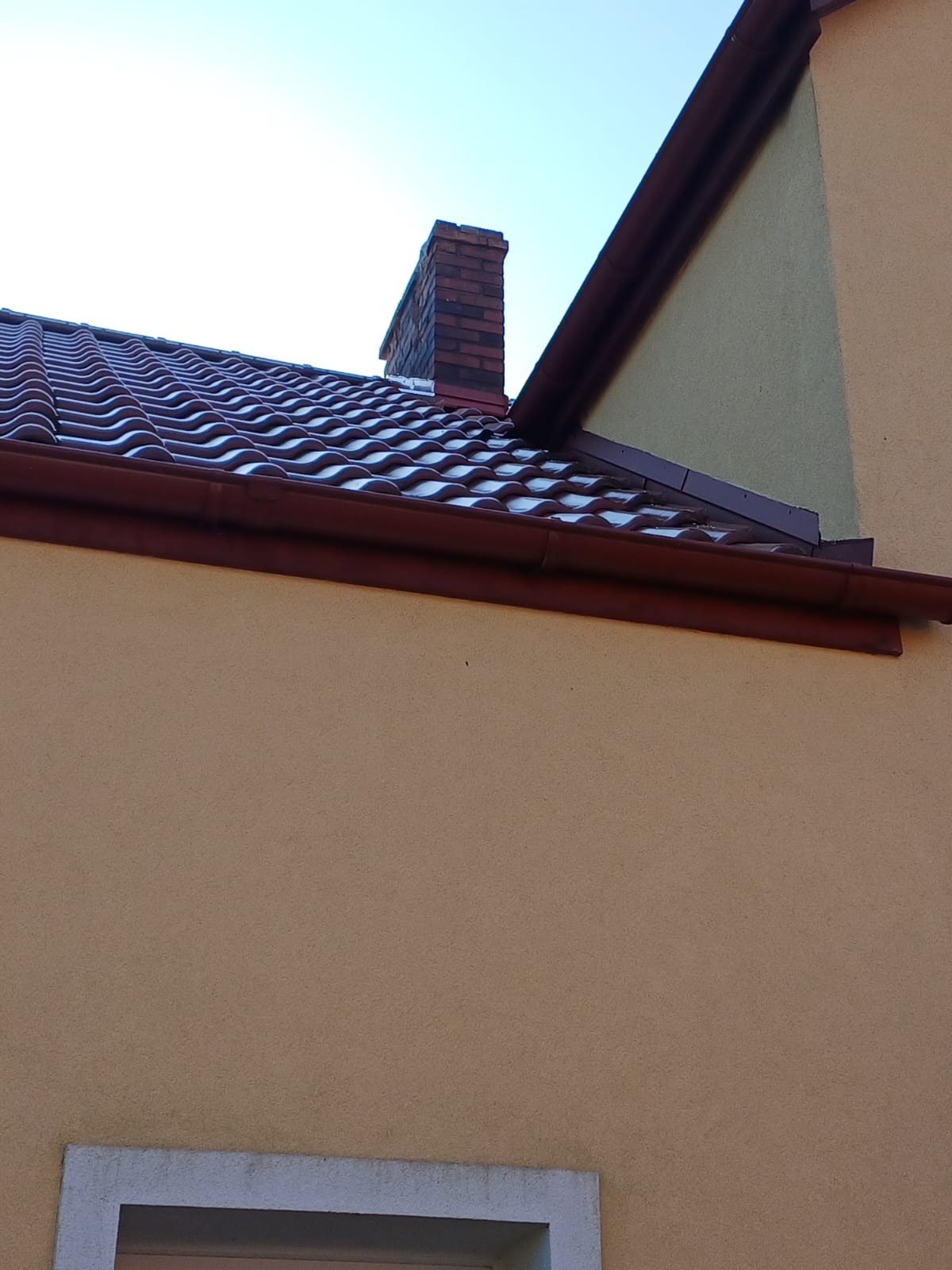 Nowy dach z brązowej dachówki ceramicznej i orynnowaniem, widoczny komin z cegły na tle błękitnego nieba, fragment elewacji budynku w kolorze piaskowym.