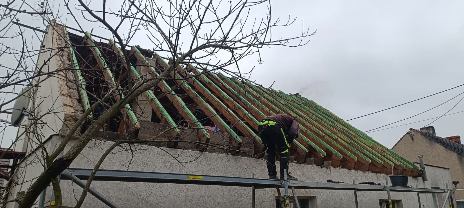 Prace dekarskie: dekarz na rusztowaniu montuje drewnianą konstrukcję dachu z zielonymi elementami wzmacniającymi. Widoczne uszkodzenia ściany szczytowej.