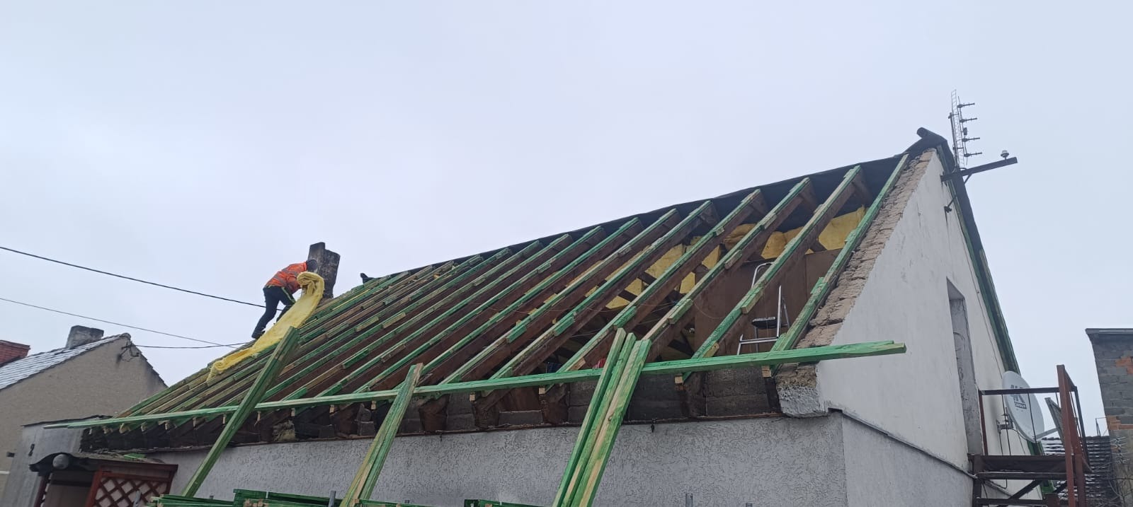 Montaż izolacji na drewnianej konstrukcji dachu domu jednorodzinnego. Widoczny pracownik w pomarańczowej kamizelce. W tle pochmurne niebo.