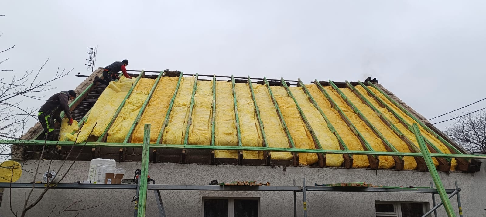 Ocieplanie dachu wełną mineralną, widoczne krokwie i pracownicy w trakcie montażu. Ujęcie z dołu, widoczne rusztowanie i materiały budowlane.