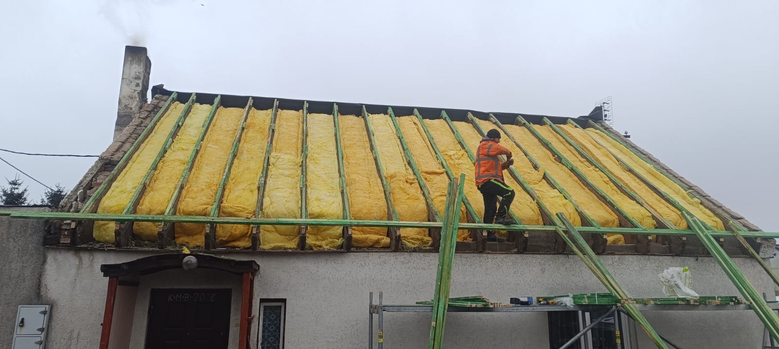 Prace dekarskie: montaż izolacji z wełny mineralnej na konstrukcji dachu domu jednorodzinnego. Widoczny dekarz w odblaskowej kurtce podczas pracy.