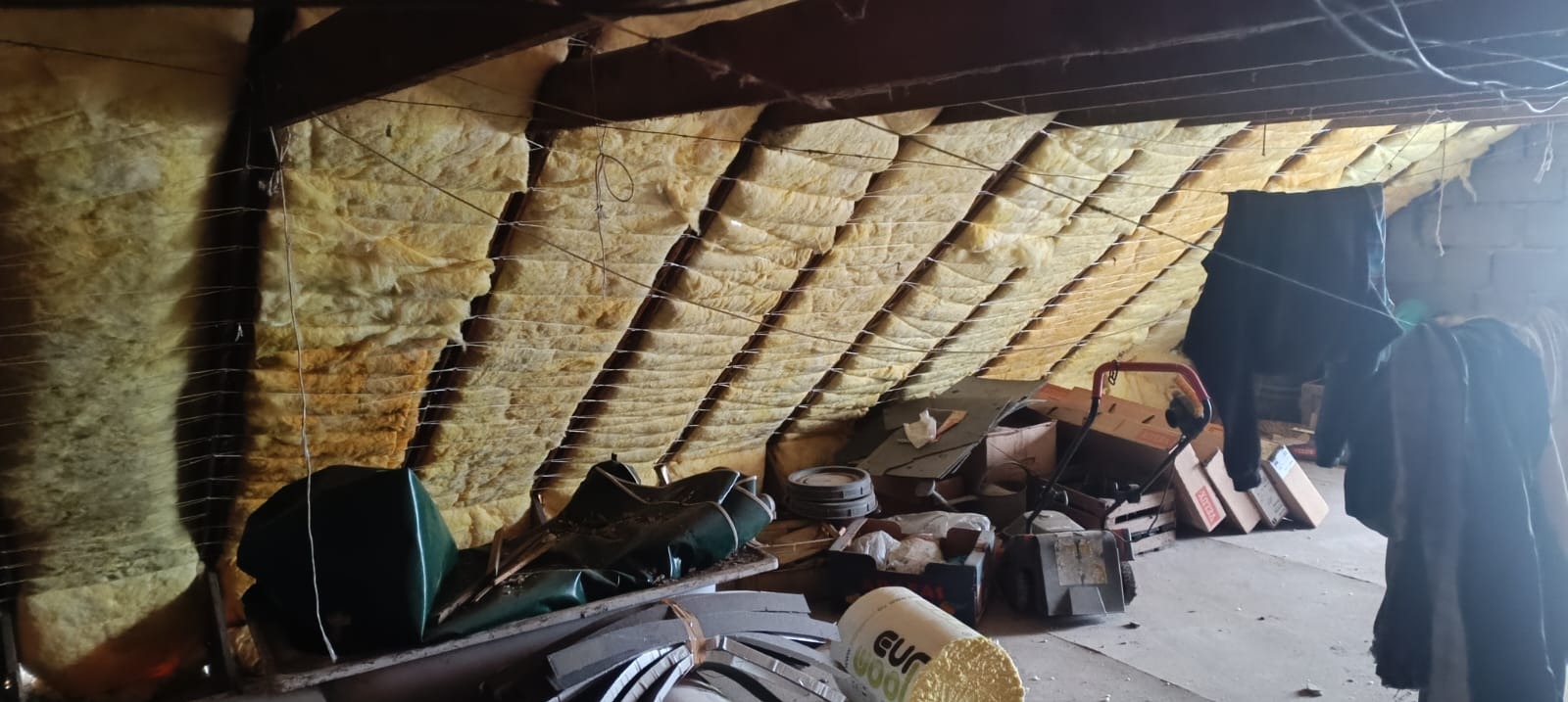 Ocieplenie poddasza wełną mineralną, widoczne mocowania z drutu. Na podłodze zgromadzone różne przedmioty, w tle ubrania na sznurku.