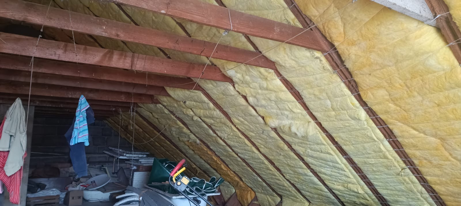 Ocieplenie poddasza wełną mineralną, widoczne drewniane krokwie i zabezpieczająca siatka. W tle nieporządek i wiszące ubrania.