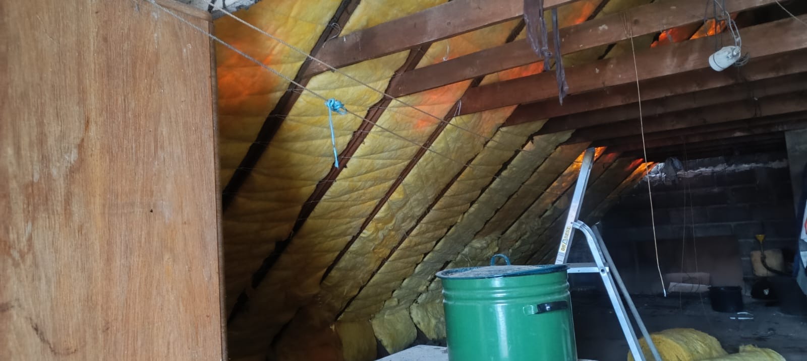 Ocieplenie poddasza wełną mineralną, widoczne krokwie i elementy konstrukcji dachu. Na pierwszym planie zielony pojemnik i drabina. Prace dekarskie w trakcie realizacji.